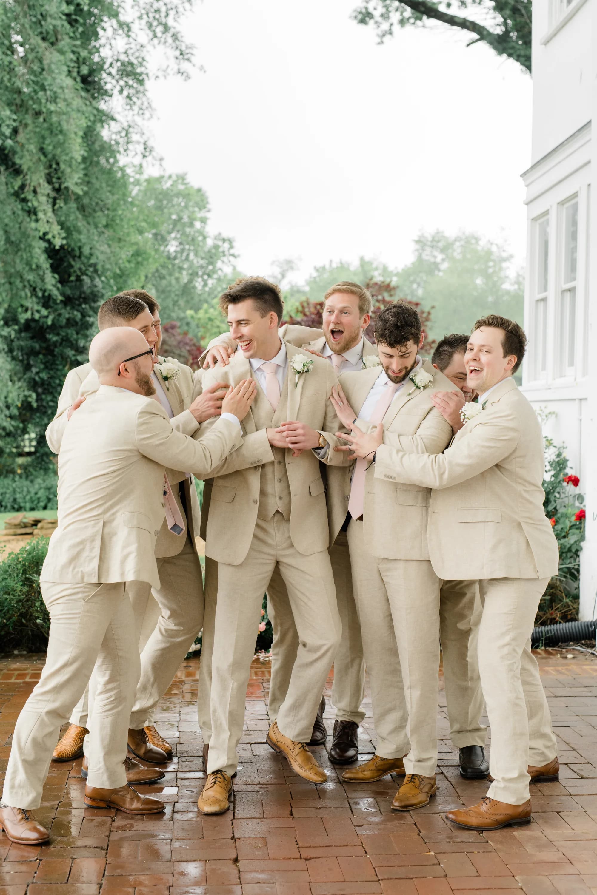 Groom and groomsmen in tan suits playfully posing together at Rixey Manor, white building visible