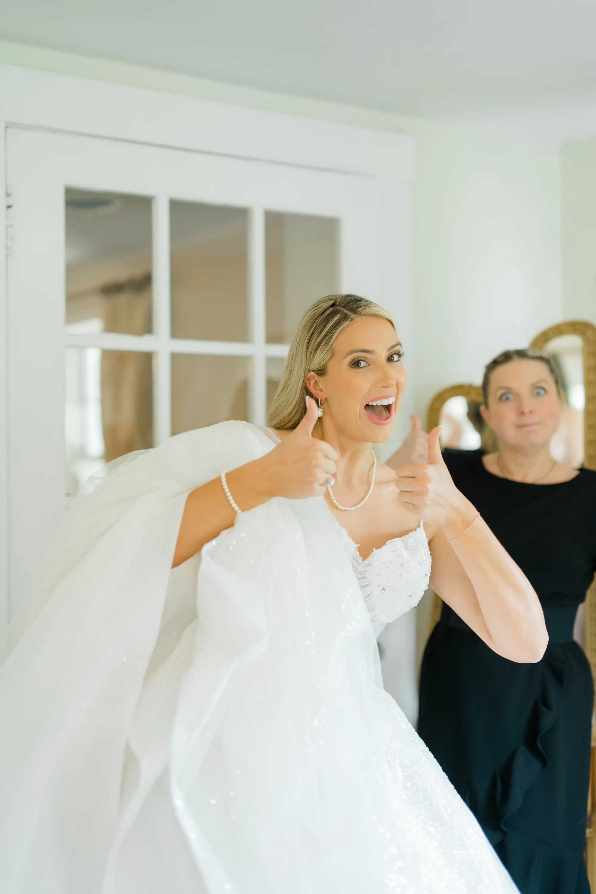 Excited bride in white gown gives two thumbs up while getting ready, helper adjusting veil behind her
