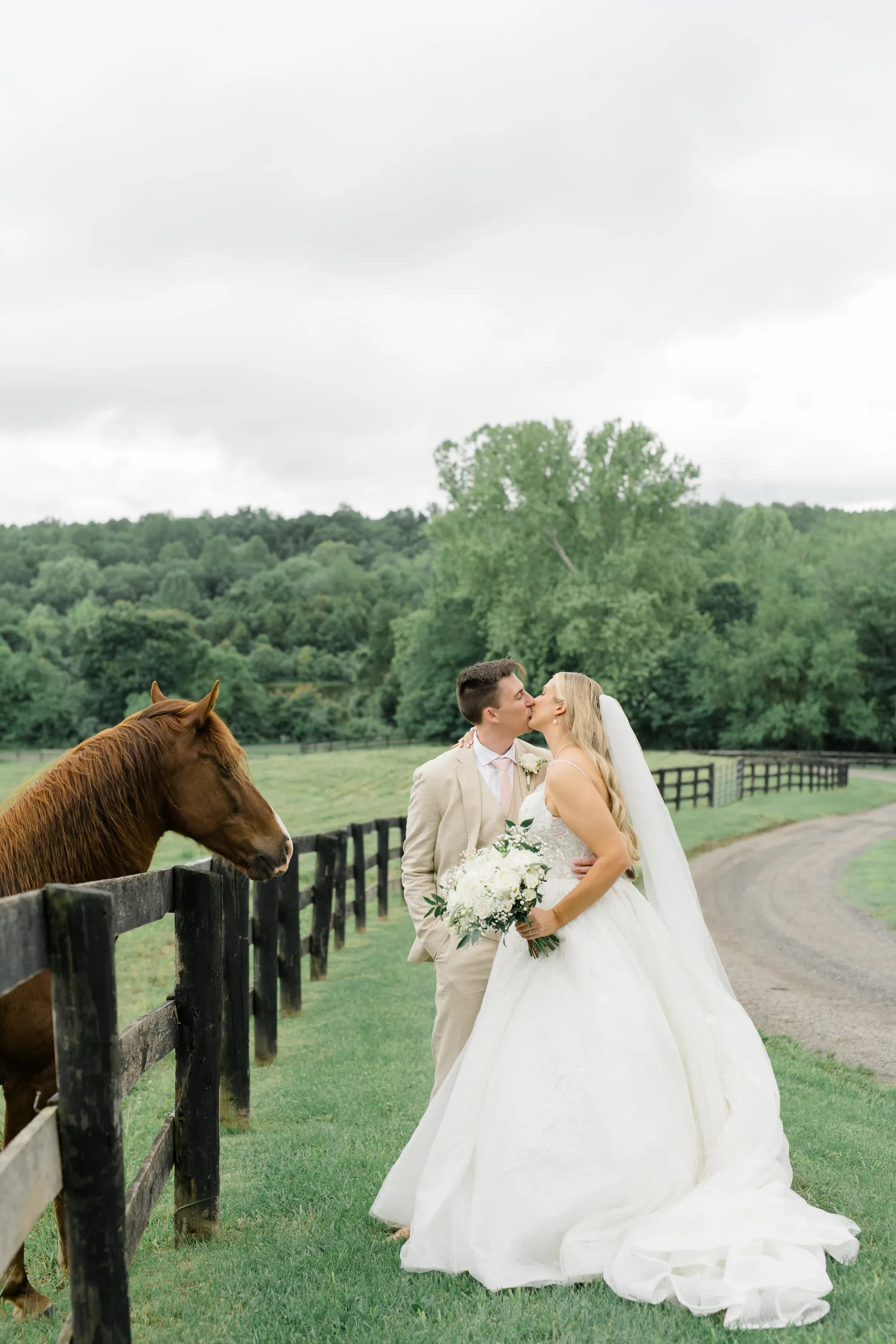 Bride and groom kiss beside a chestnut horse at a black fence-lined pasture at Rixey Manor