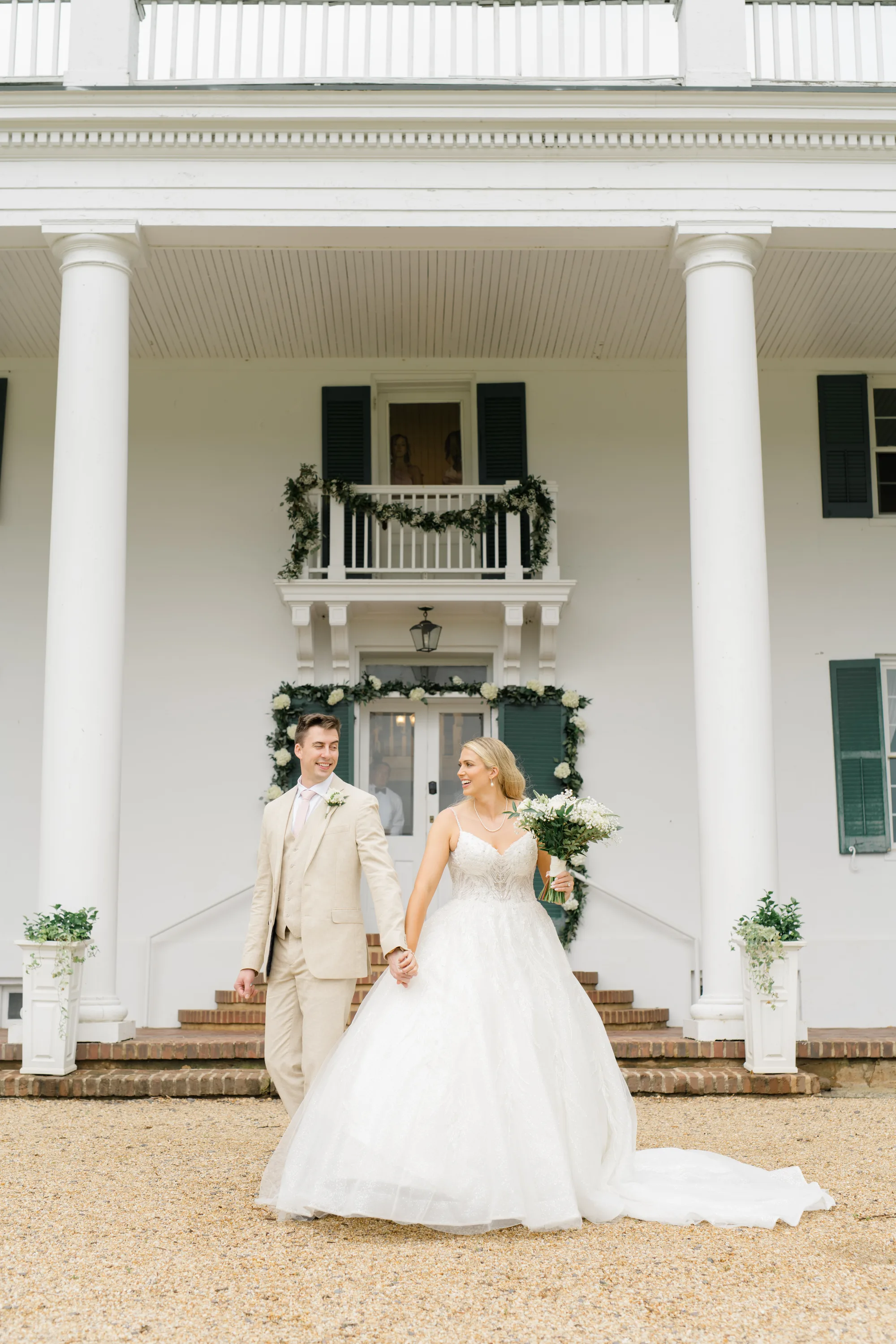 Bride and groom holding hands and smiling in front of Rixey Manor's white columned facade with floral arch