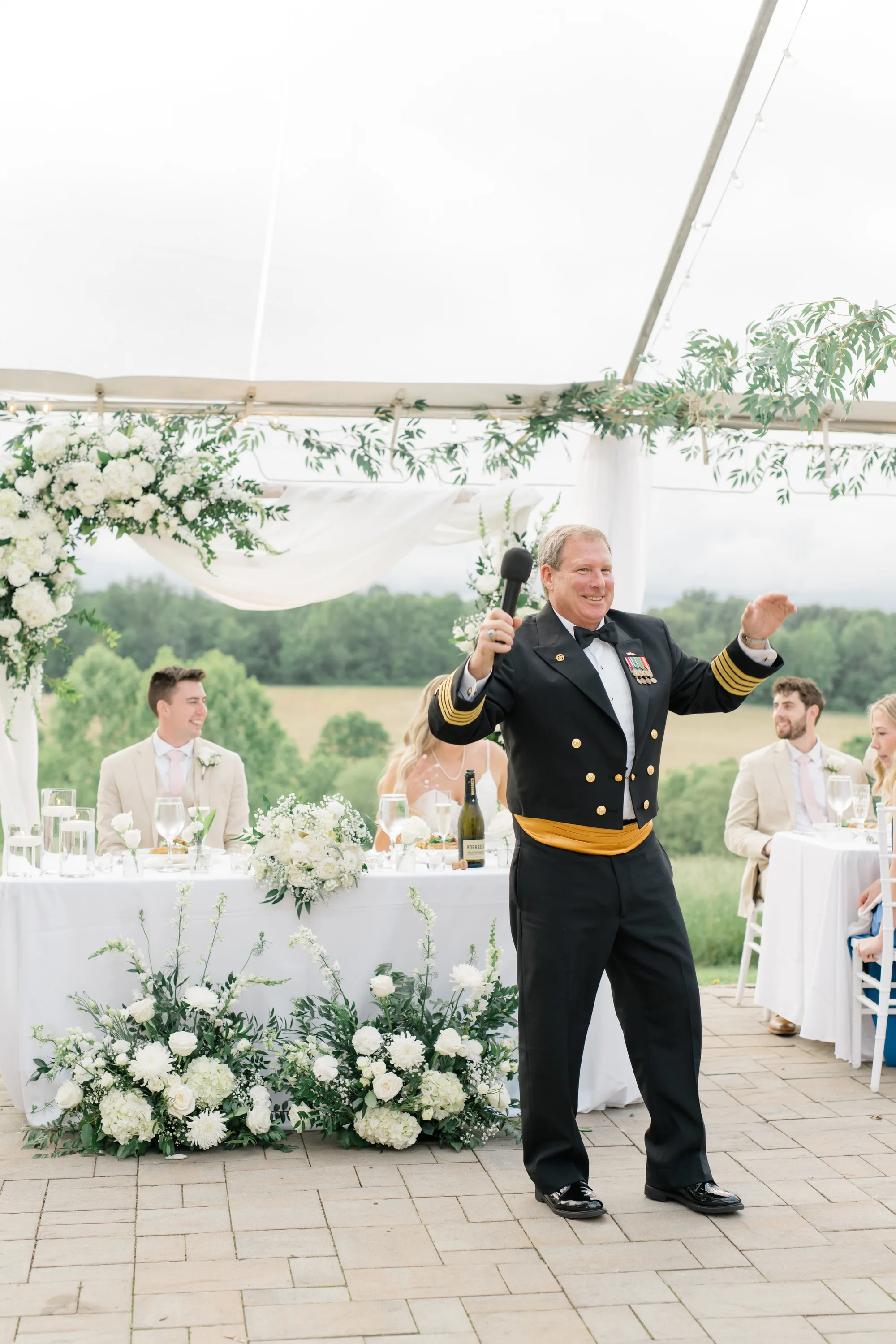 Naval officer delivers toast at Rixey Manor tented reception with white floral arrangements and pastoral Virginia views