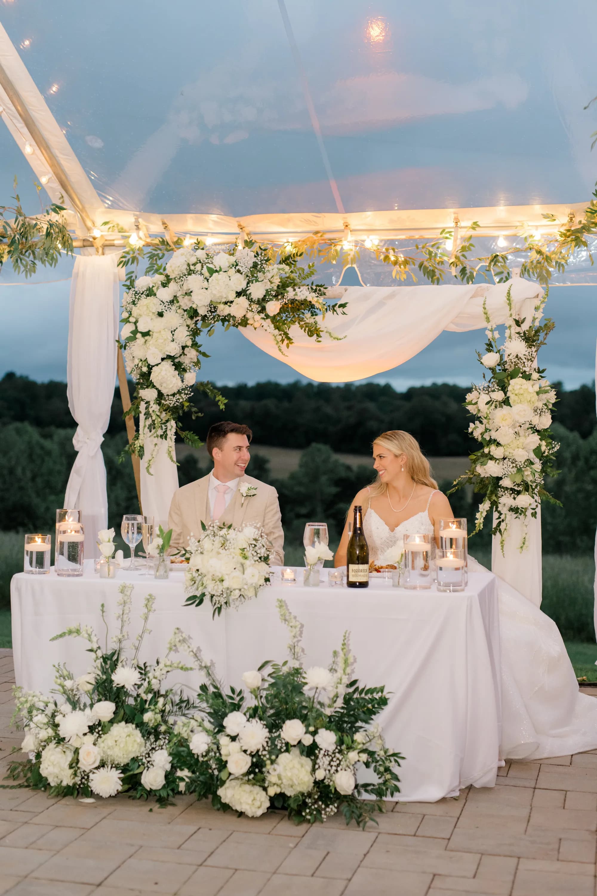 Bride and groom share a smile at sweetheart table beneath floral arch in Rixey Manor's tented reception at dusk
