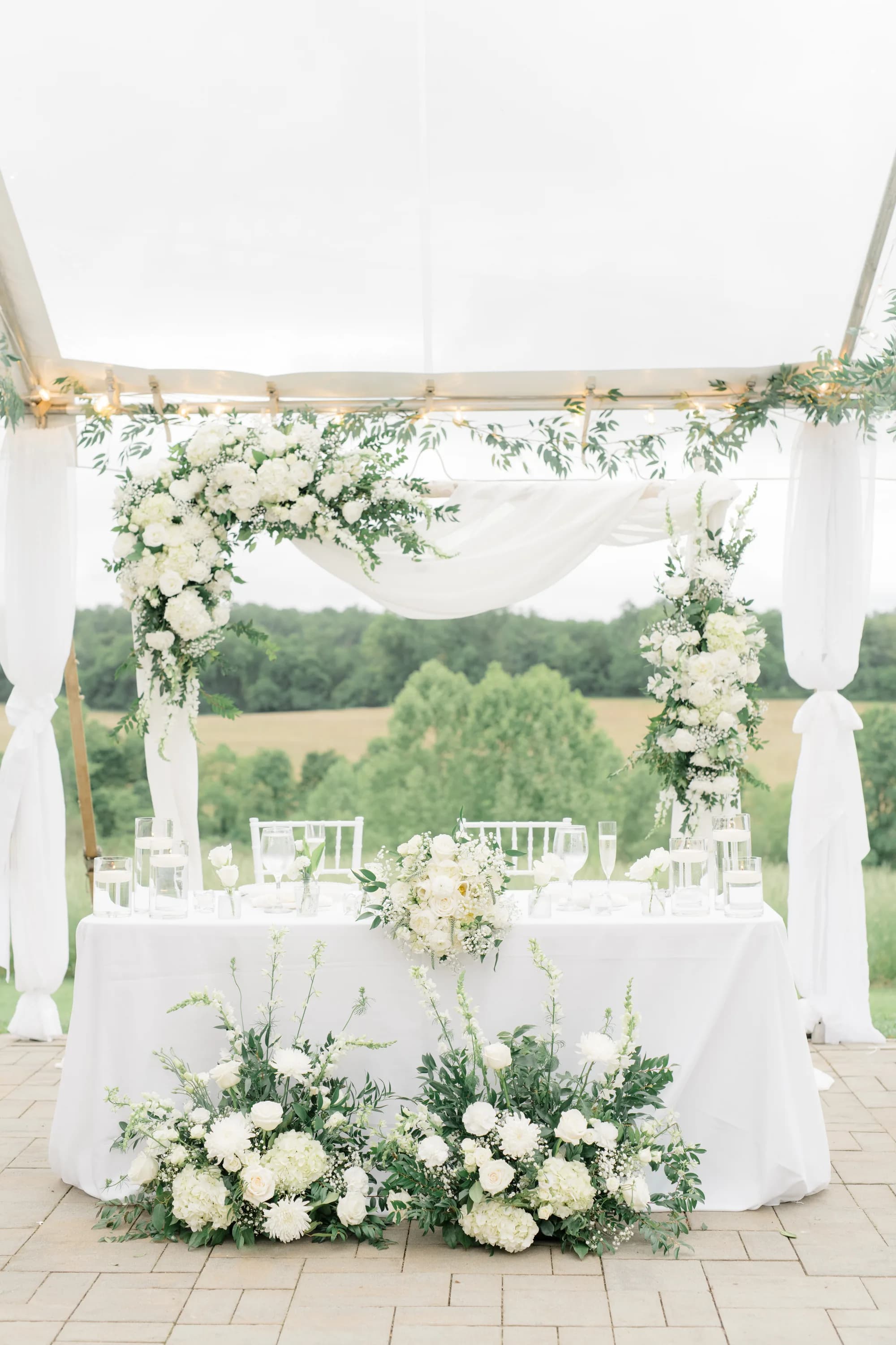 Sweetheart table adorned with white floral arches under Rixey Manor's tent with rolling Virginia countryside behind