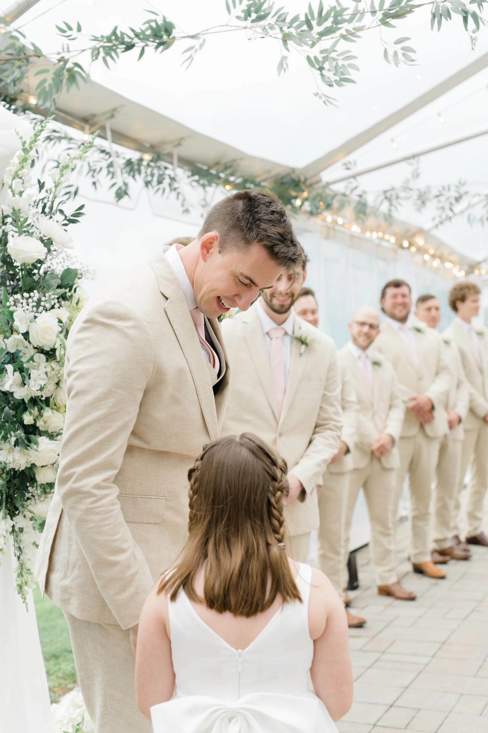 Groom smiles down at flower girl during ceremony under Rixey Manor's tent with groomsmen lined behind