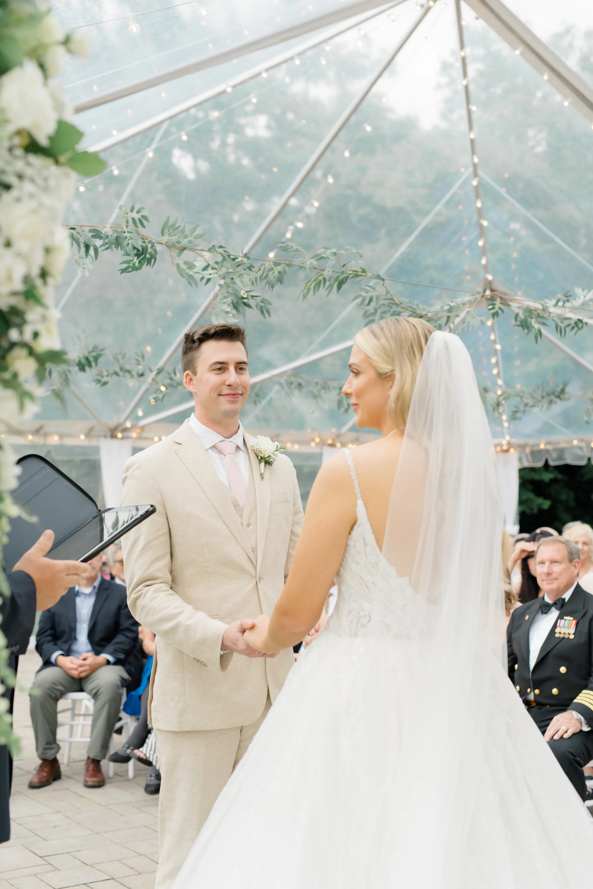 Groom smiles at bride during vows under Rixey Manor's clear tent adorned with greenery and fairy lights