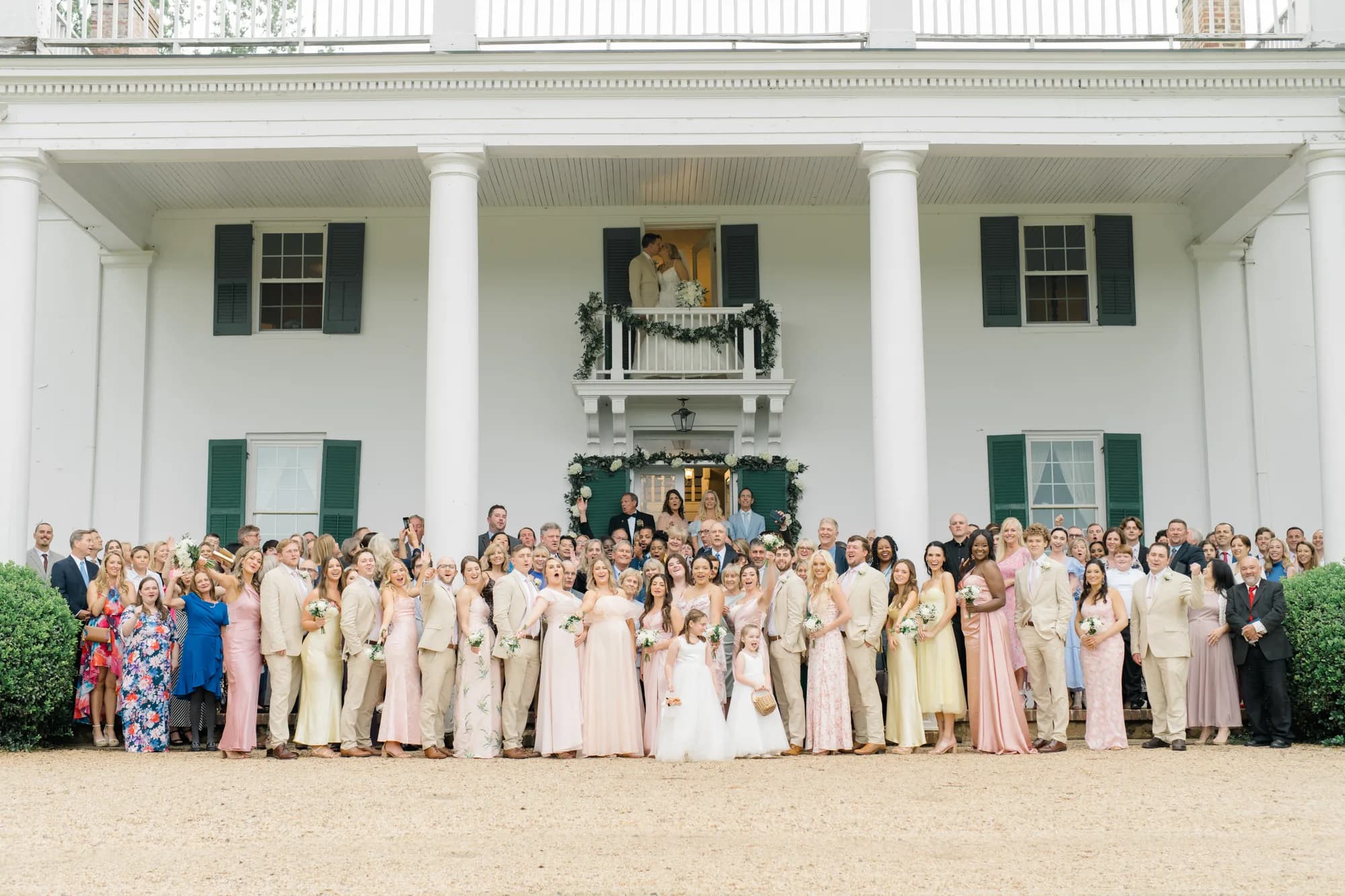 Full wedding party group portrait in front of Rixey Manor's white columned facade with couple on balcony above