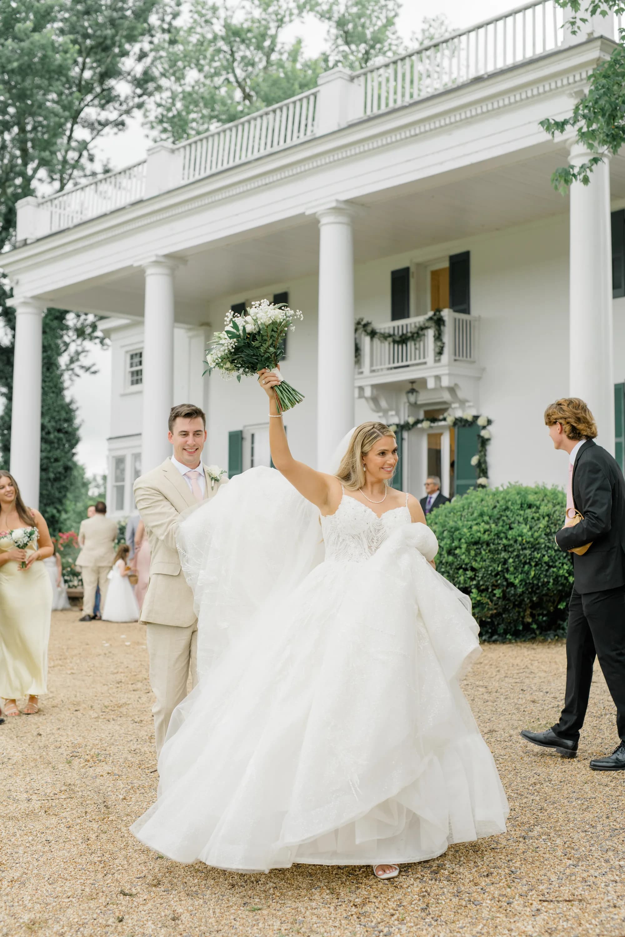 Bride in white gown being lifted by groom in tan suit, holding bouquet, with manor columns in background