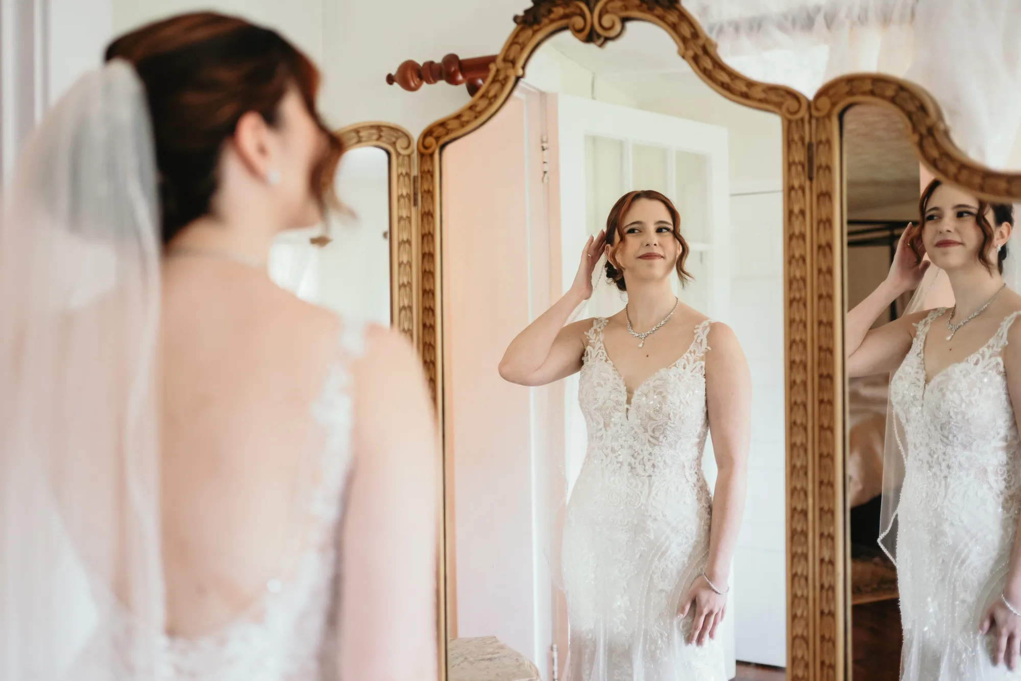 Bride in lace gown adjusting earring while admiring her reflection in an ornate gold mirror