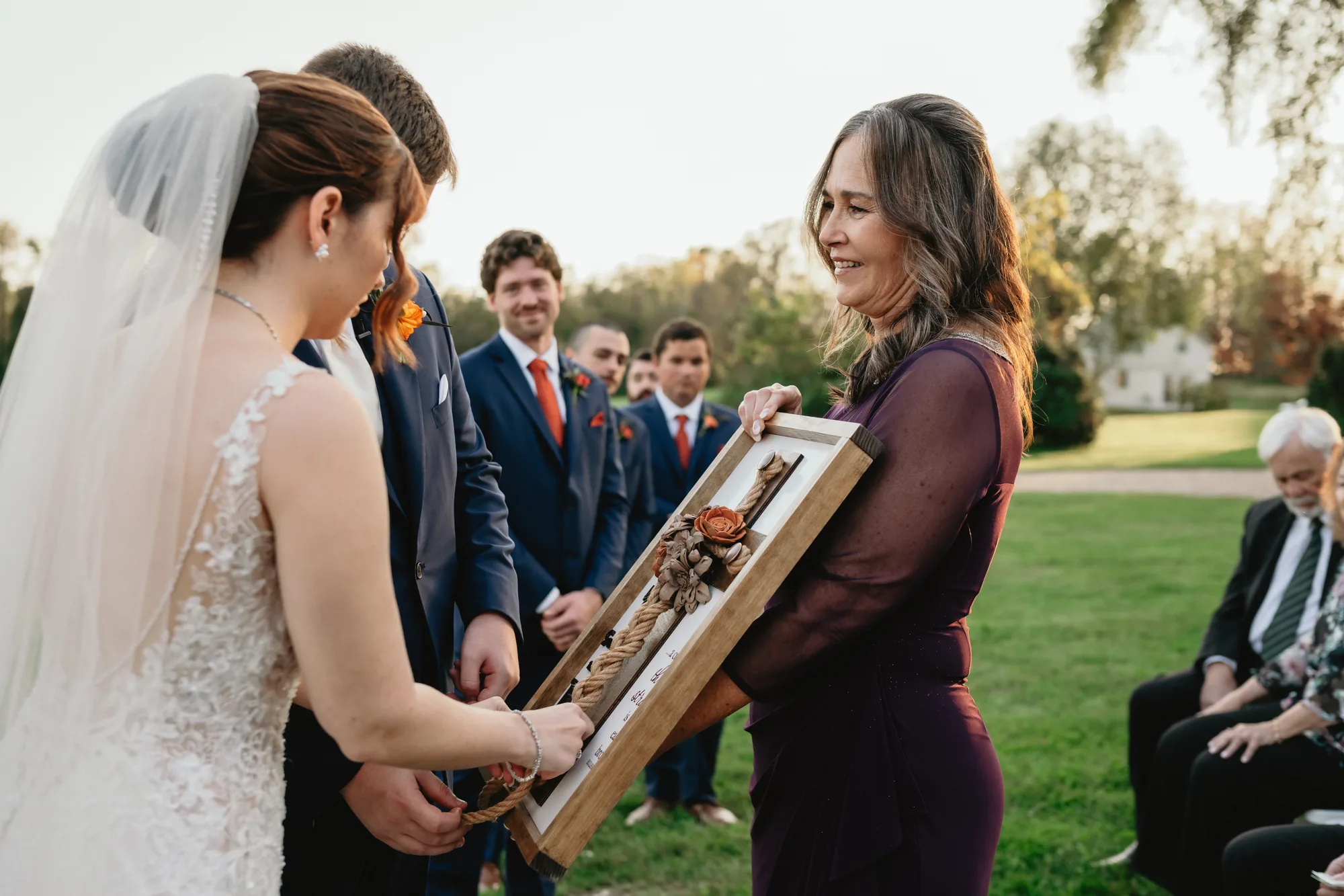 Bride receives framed wedding gift during outdoor ceremony at Rixey Manor, groomsmen and guests visible behind
