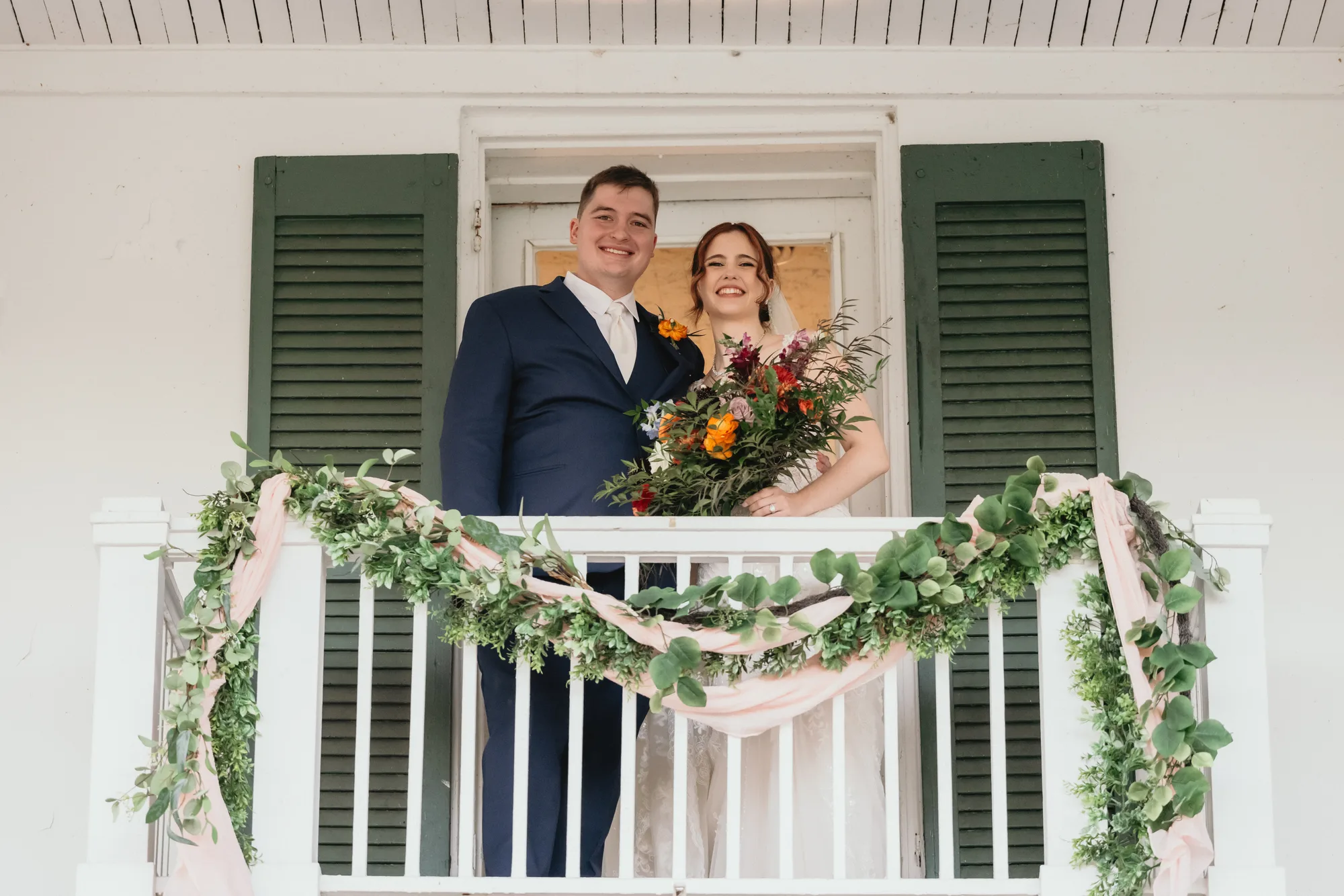 Smiling newlyweds on a garland-draped balcony at Rixey Manor, bride holding a colorful wildflower bouquet