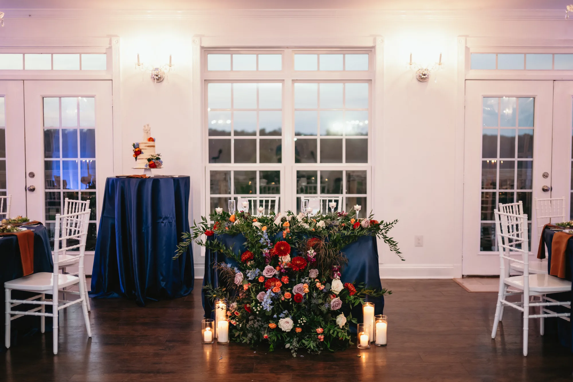 Sweetheart table with lush floral arrangement and candles in Rixey Manor ballroom, wedding cake table behind