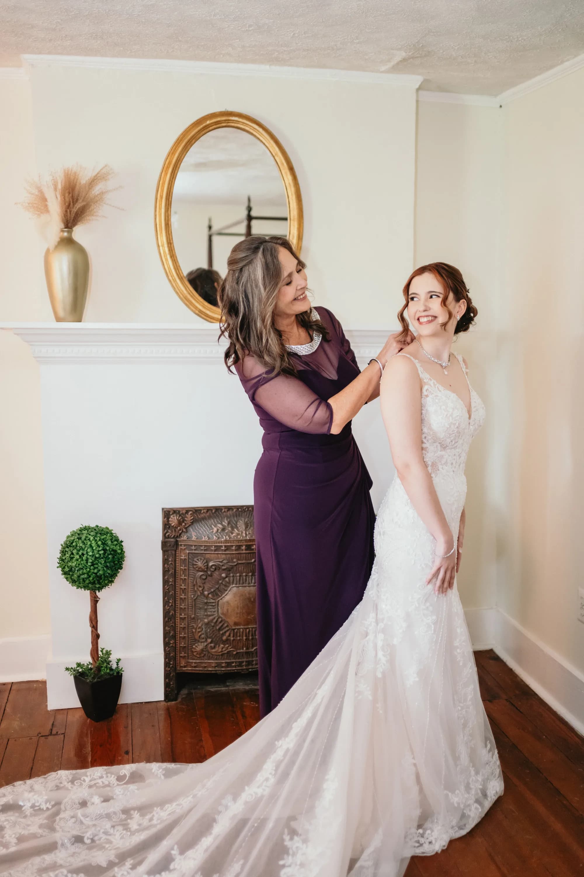 Mother helps bride with necklace before ceremony, both smiling, in a bright room with fireplace and gold mirror