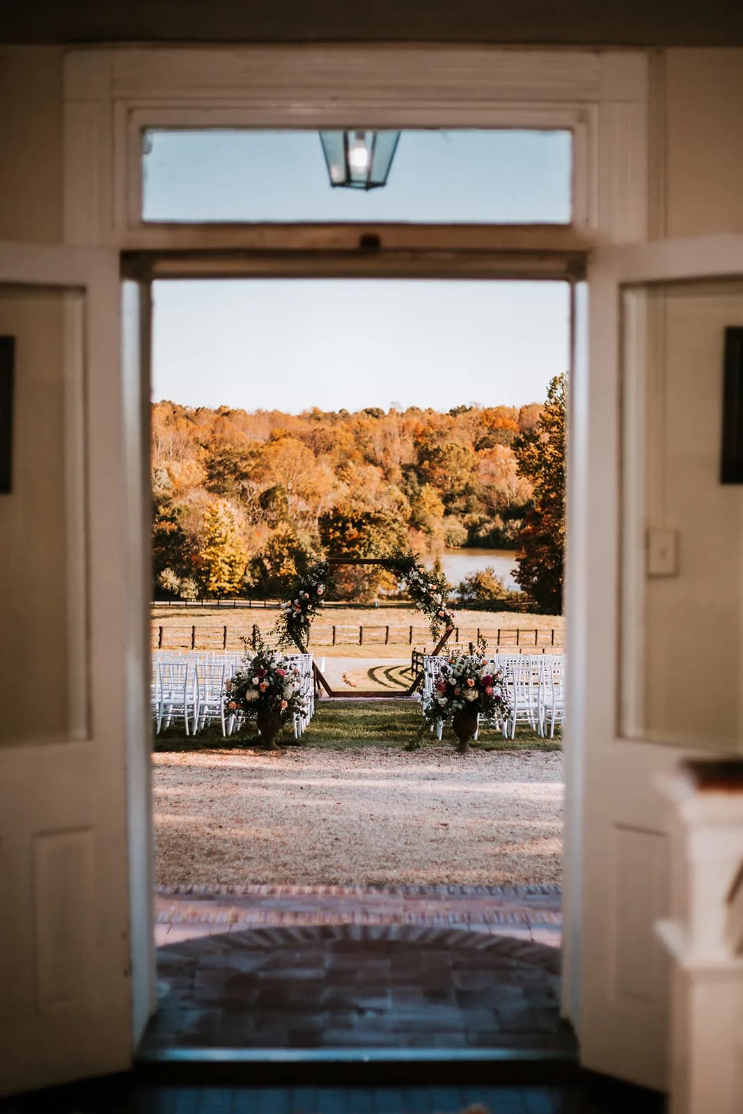 View through white manor doors of outdoor wedding ceremony setup with white chairs and floral arch by water