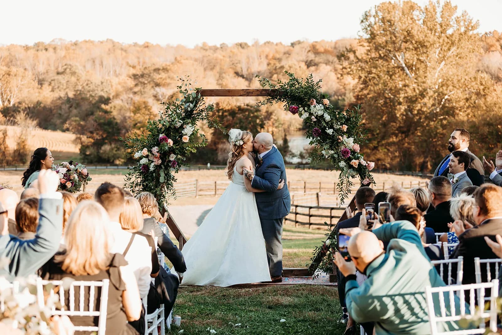 Bride and groom share first kiss under floral arch at Rixey Manor outdoor ceremony, guests cheering in golden autumn light
