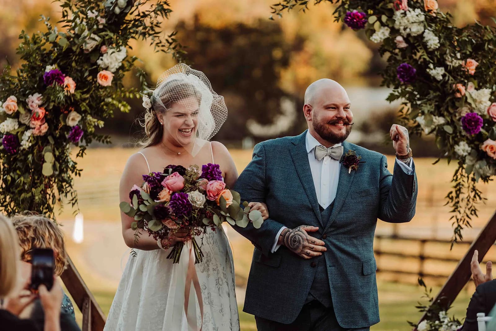 Joyful newlyweds celebrate under floral arch at Rixey Manor outdoor ceremony, groom pumping fist in excitement