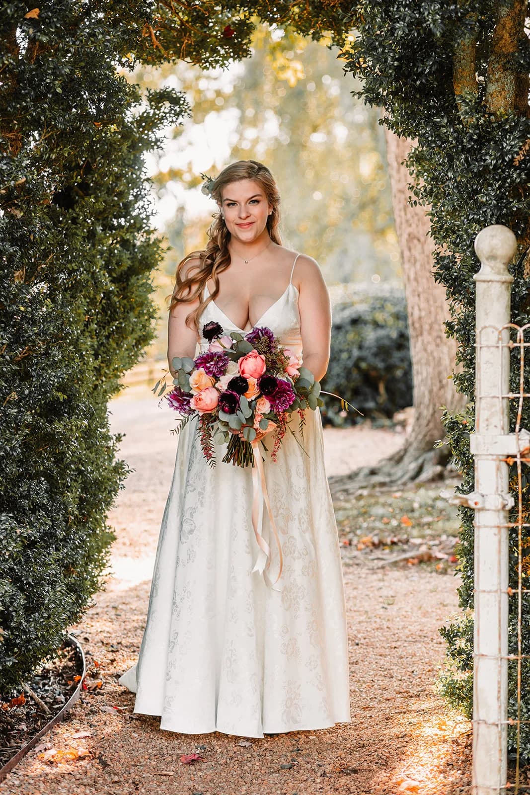 Bride in floral gown holding jewel-toned bouquet beneath a lush garden archway at Rixey Manor