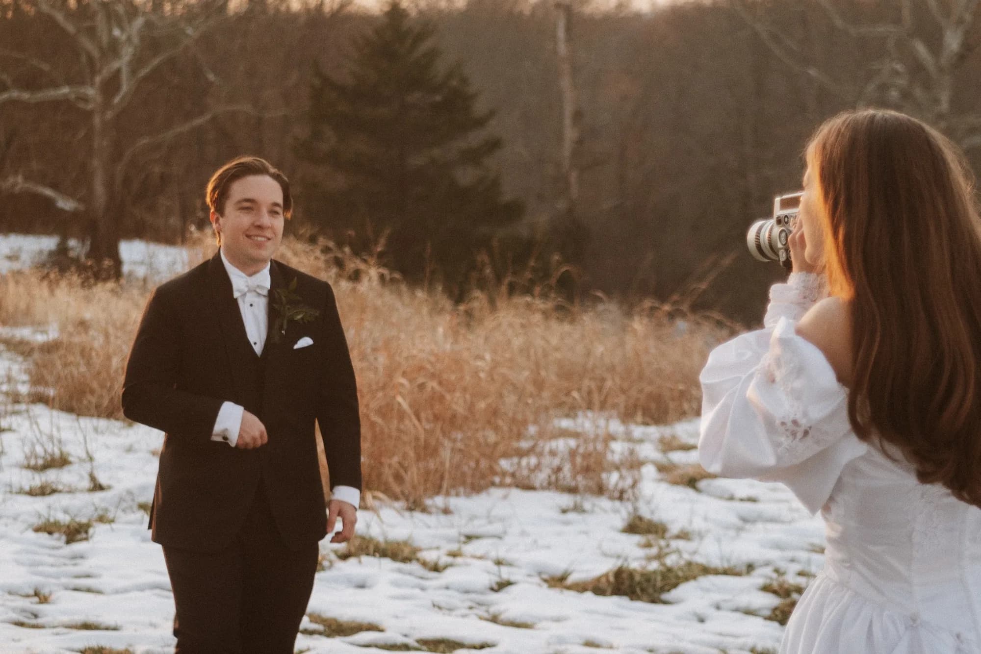 Bride photographs groom in snowy field at golden hour during winter wedding portrait session