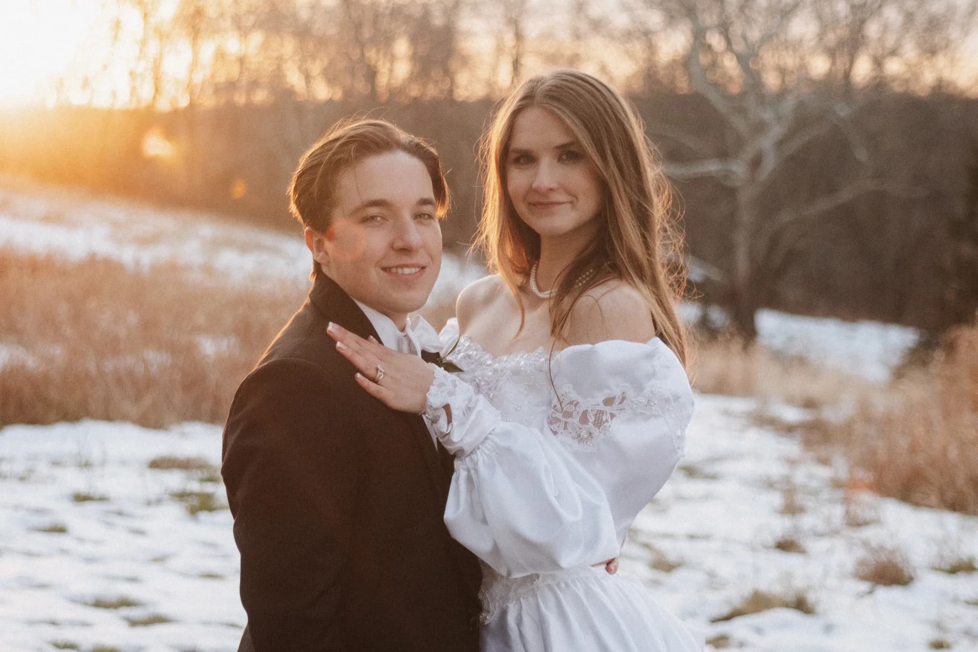 Bride and groom embrace in a snowy field at golden hour, bride in off-shoulder white gown