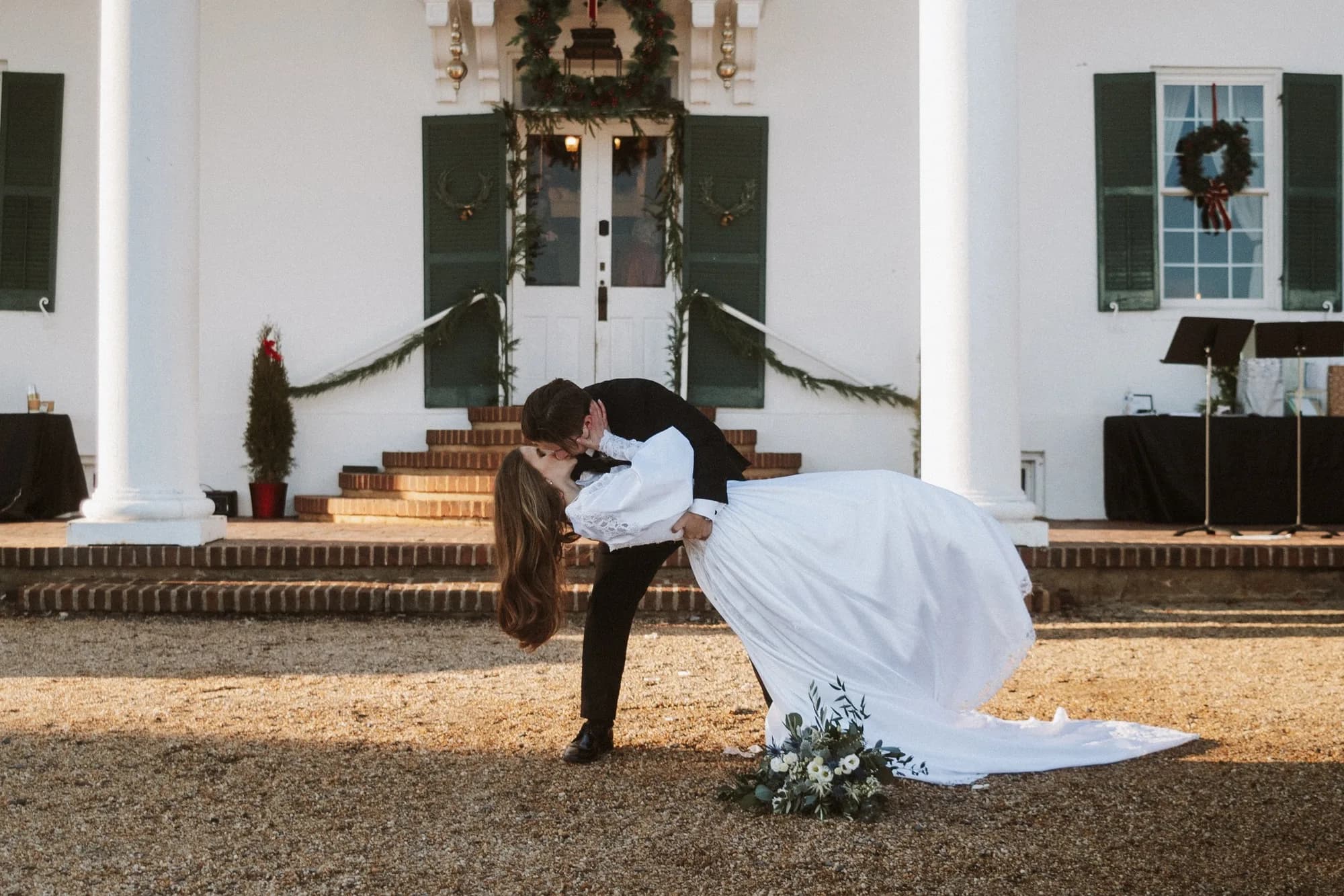 Groom dips bride in a romantic kiss before Rixey Manor's white columned entrance adorned with Christmas greenery