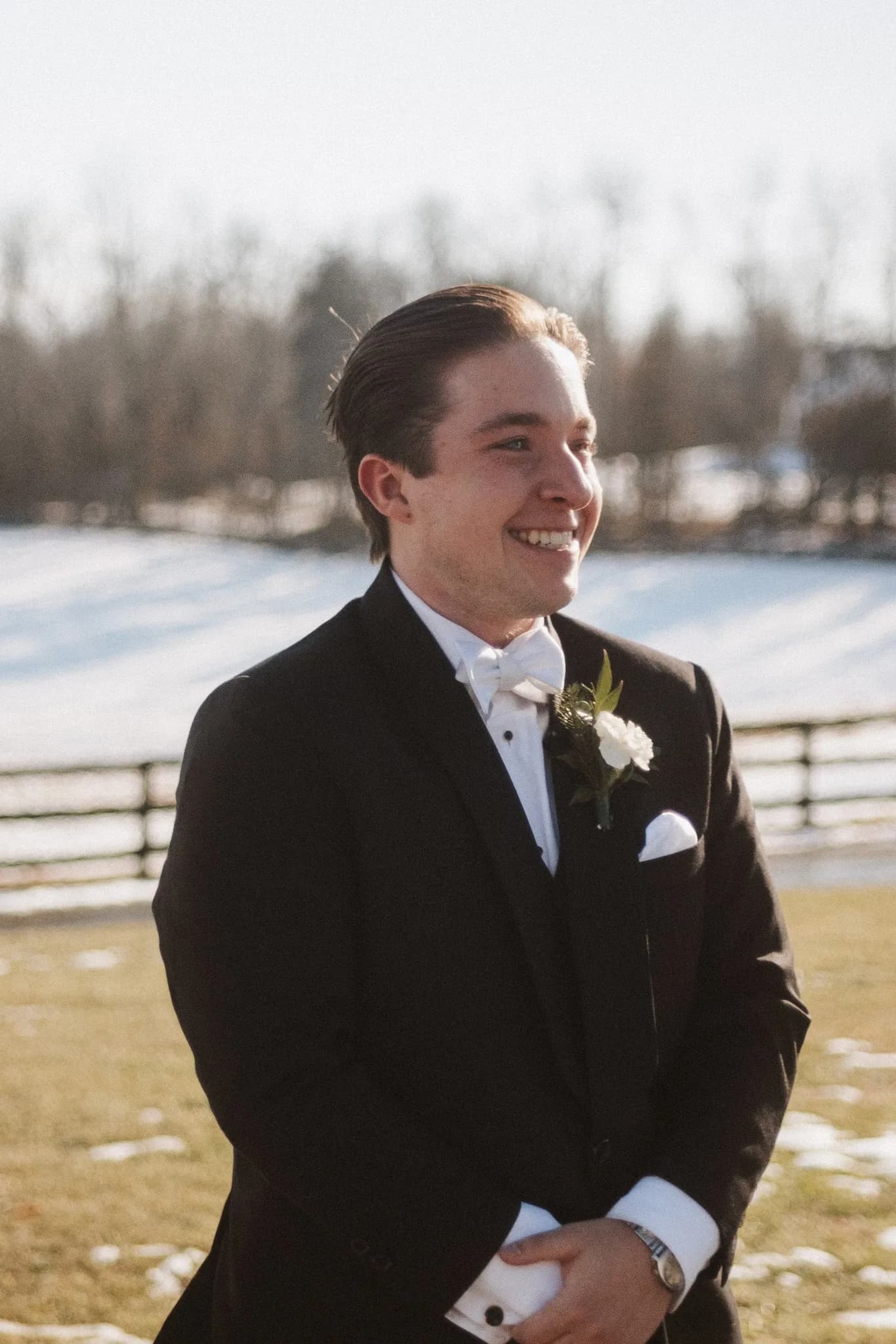 Groom smiling joyfully outdoors in black tuxedo with white boutonniere against snowy Virginia field