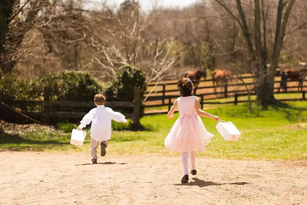 Two children in wedding attire running across sunny Rixey Manor grounds with wooden fence and horses in background