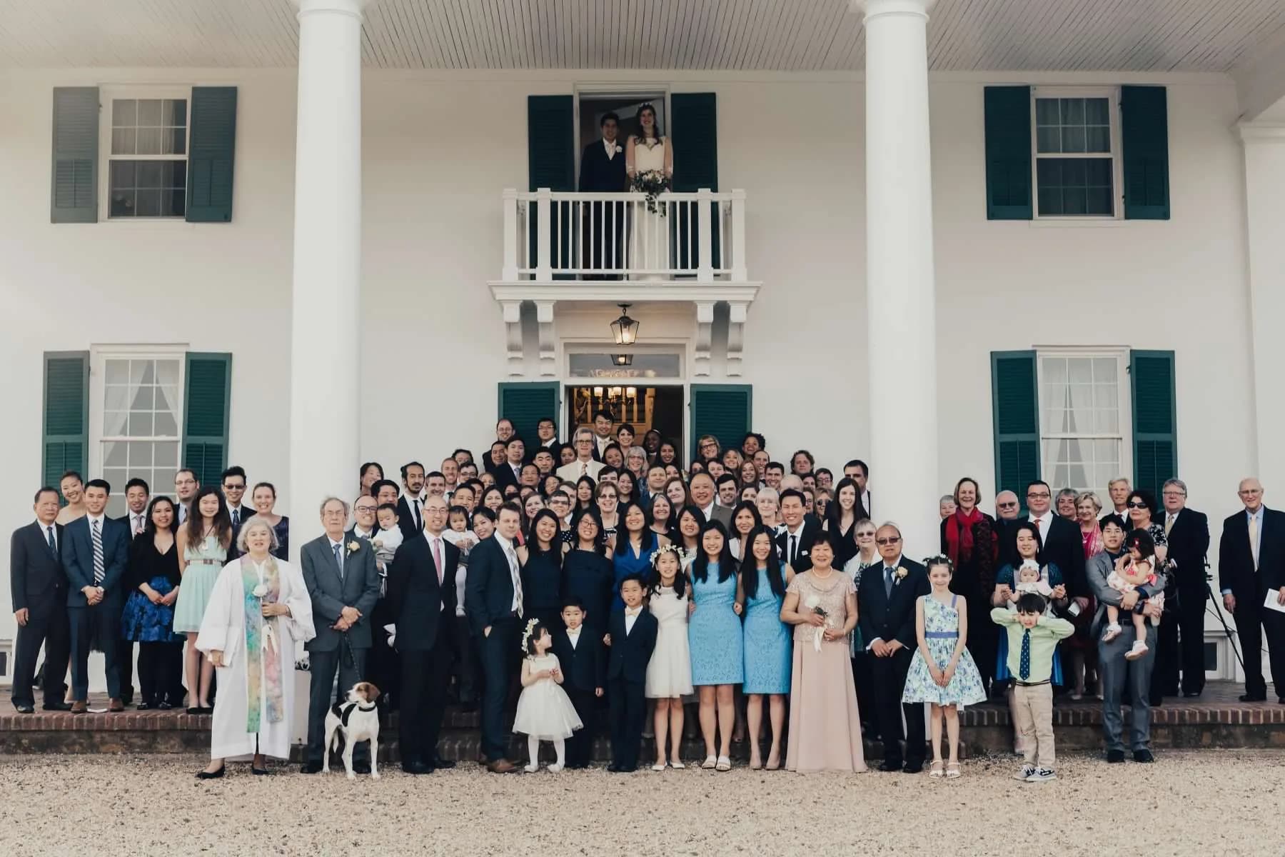 Large wedding group portrait on steps of Rixey Manor colonial estate, couple visible on upper balcony