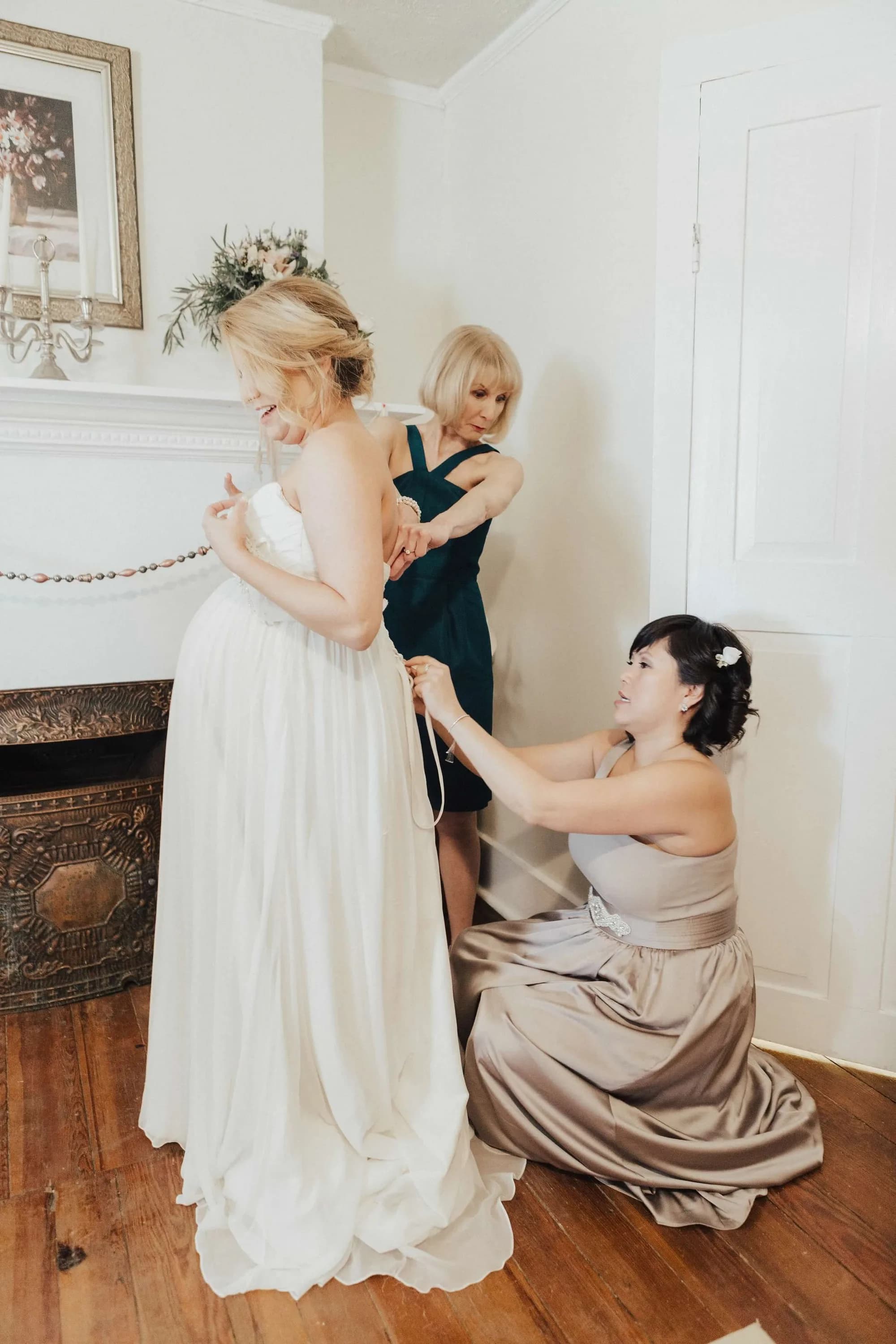 Bride smiles as mother and bridesmaid help lace her wedding gown before the ceremony