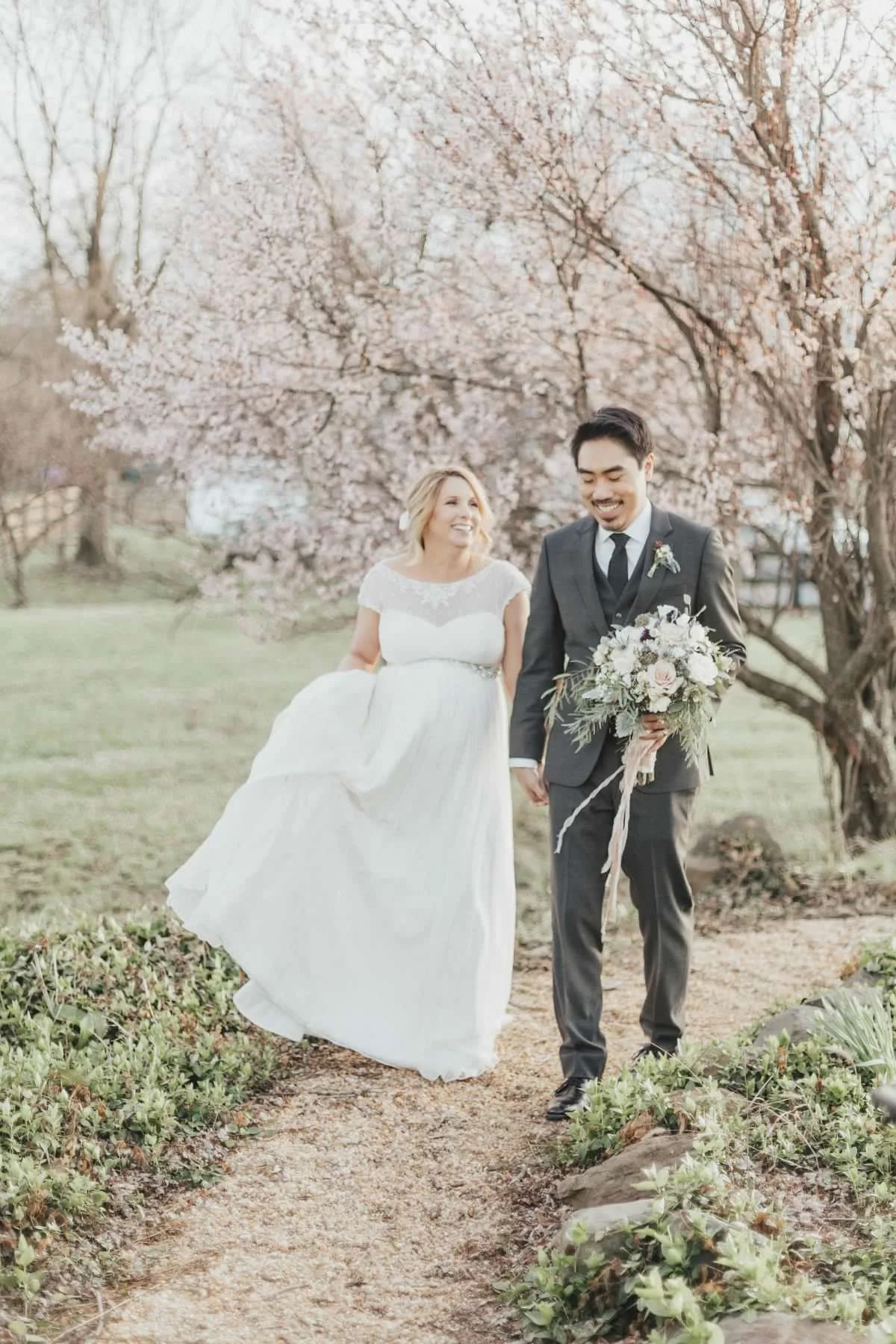 Bride and groom walk hand-in-hand along a garden path beneath blooming cherry blossom trees, both smiling