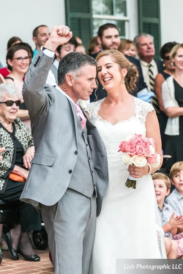 Groom pumps fist in celebration as bride laughs joyfully after wedding ceremony, guests cheering behind them
