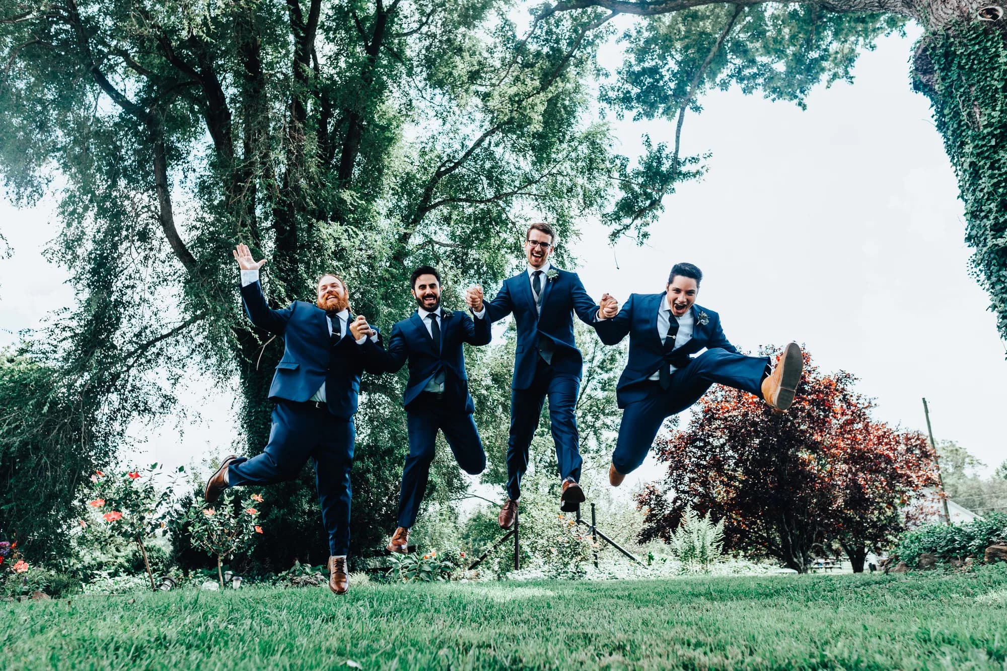 Four groomsmen in navy suits jumping joyfully on lush green lawn surrounded by trees