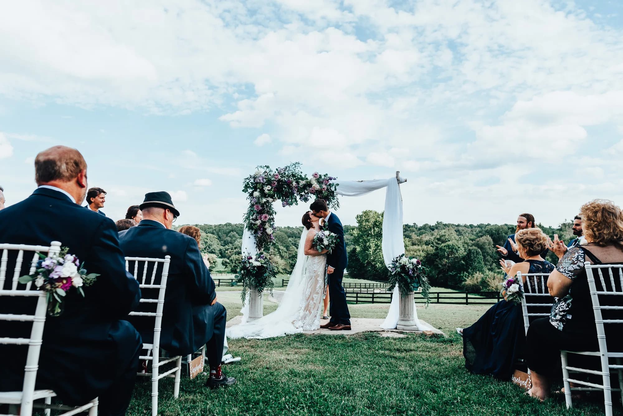 Couple shares first kiss under floral arch at Rixey Manor outdoor ceremony with rolling Virginia countryside behind them
