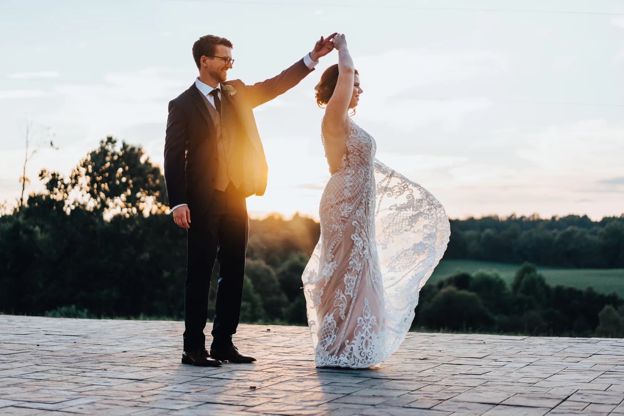 Groom twirls bride in lace gown on stone terrace at golden hour, rolling Virginia countryside in background
