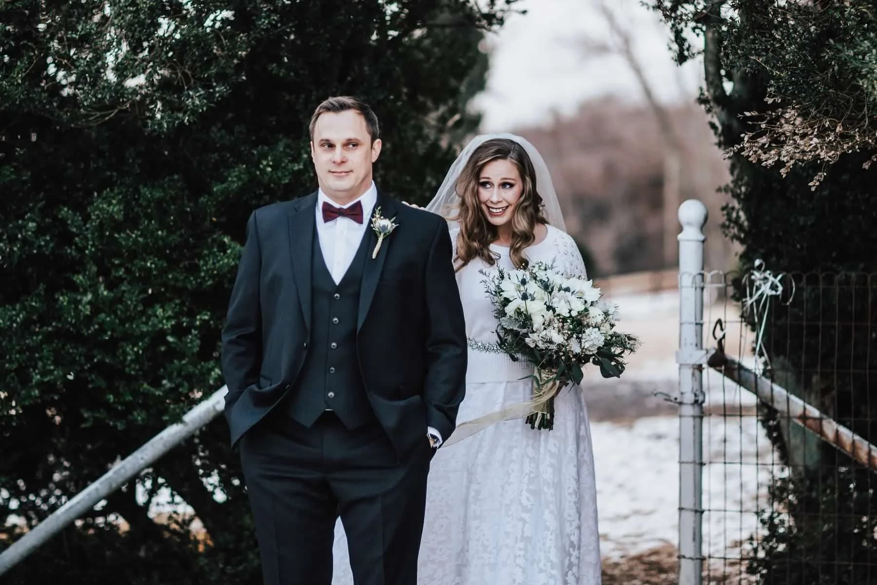 Bride and groom portrait on snowy winter grounds, bride holding white bouquet and smiling warmly