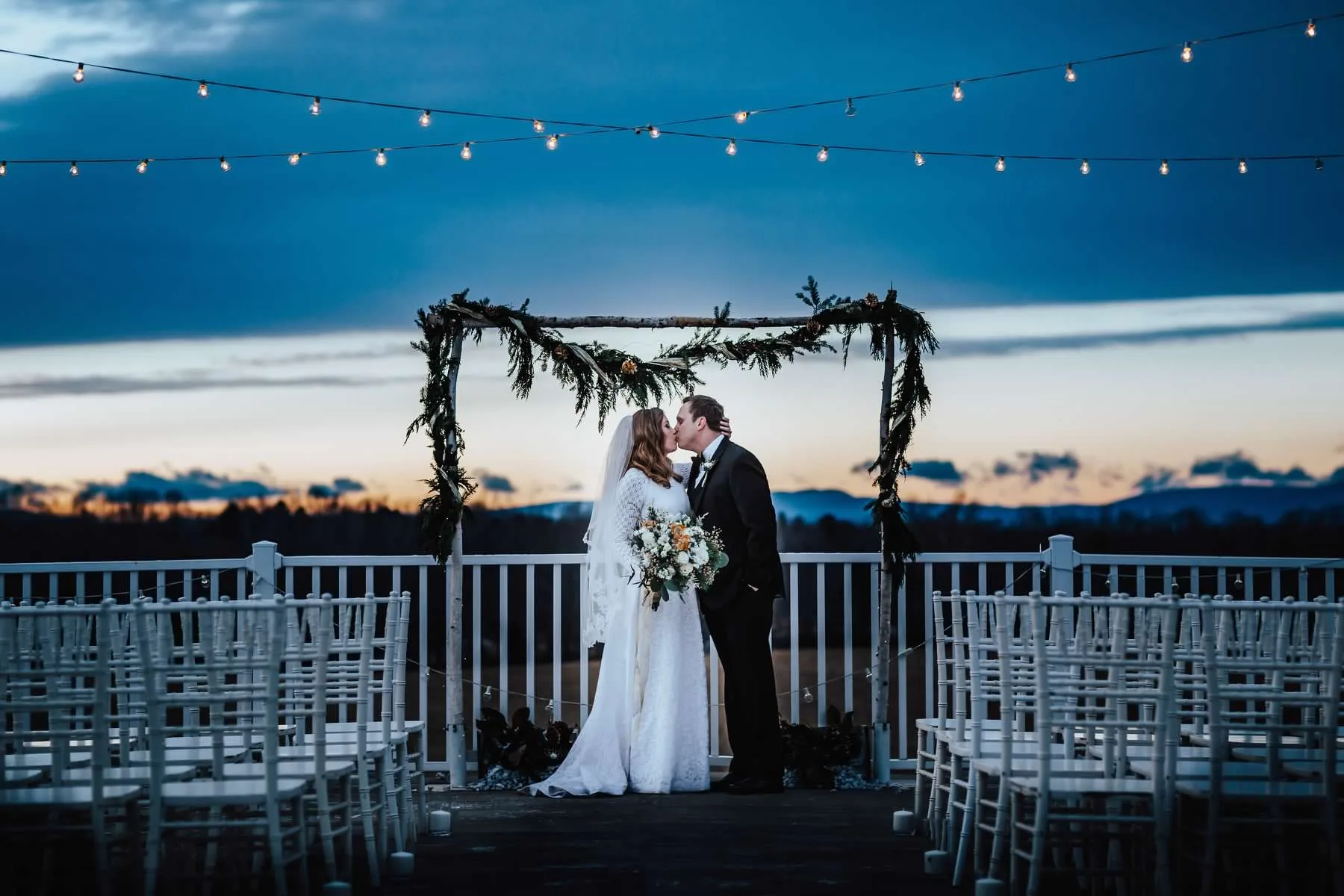 Bride and groom share first kiss under floral arch on Rixey Manor rooftop terrace at twilight with string lights