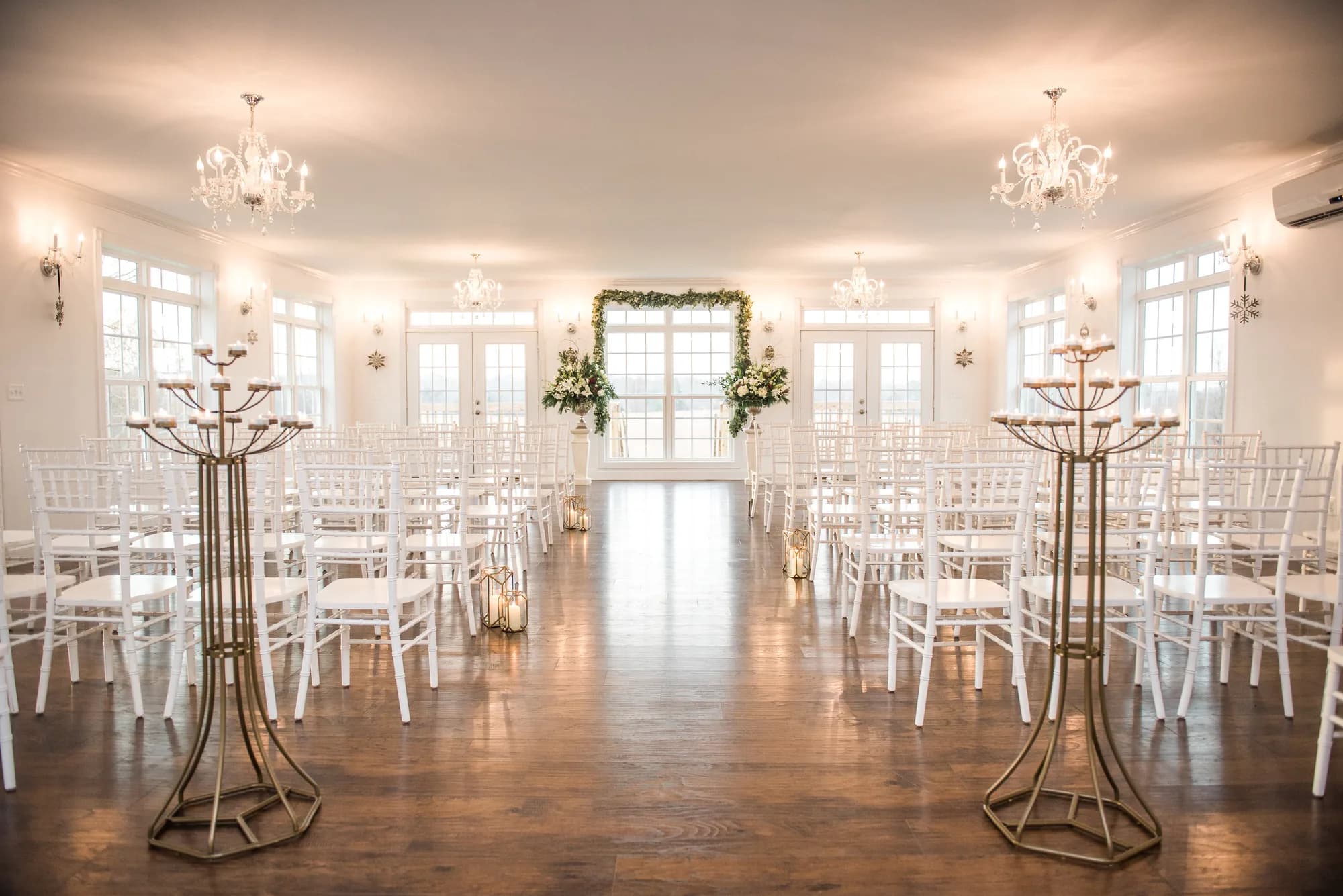 Sunlit ceremony hall at Rixey Manor with white chiavari chairs, crystal chandeliers, and floral arch at the altar