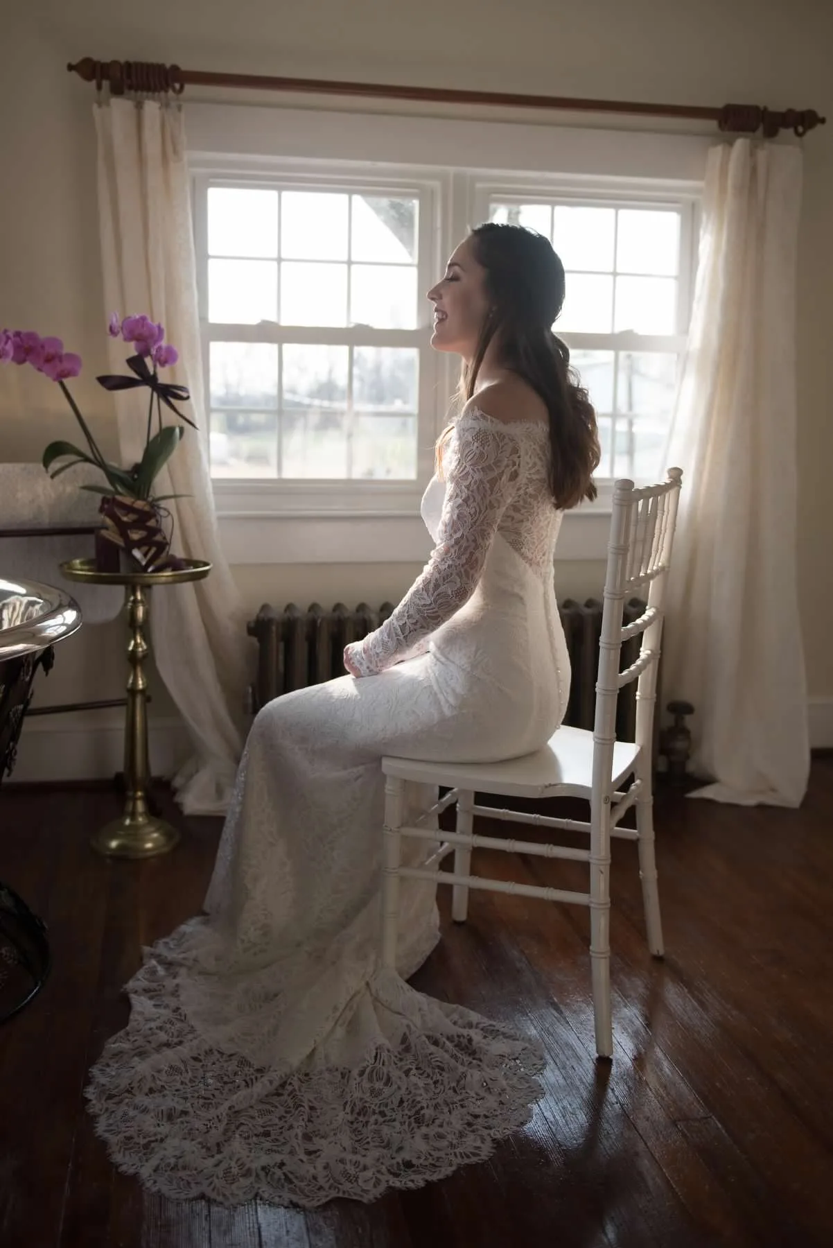 Bride in lace gown seated by sunlit window, laughing softly in a warmly lit room at Rixey Manor