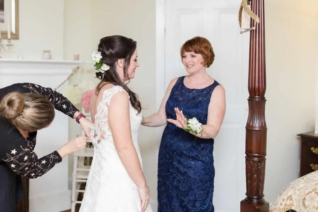 Bride in lace gown being buttoned while sharing a joyful moment with a woman in navy lace dress