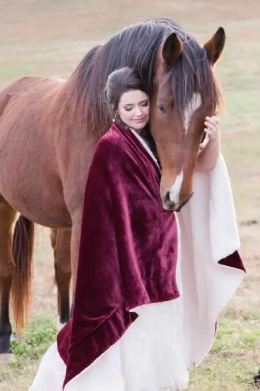 Bride in burgundy velvet wrap tenderly resting her head against a chestnut horse in a grassy field