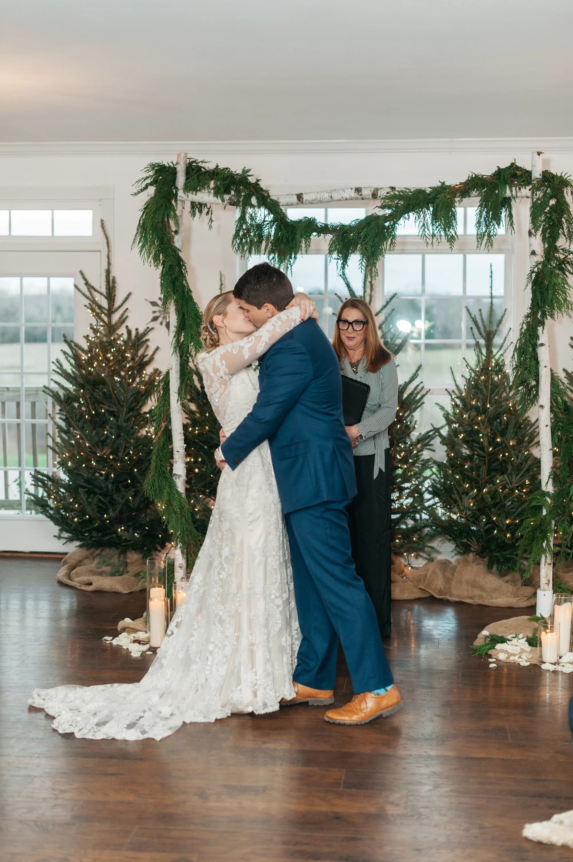 Bride and groom share first kiss at indoor winter ceremony altar adorned with evergreen garland and lit Christmas trees
