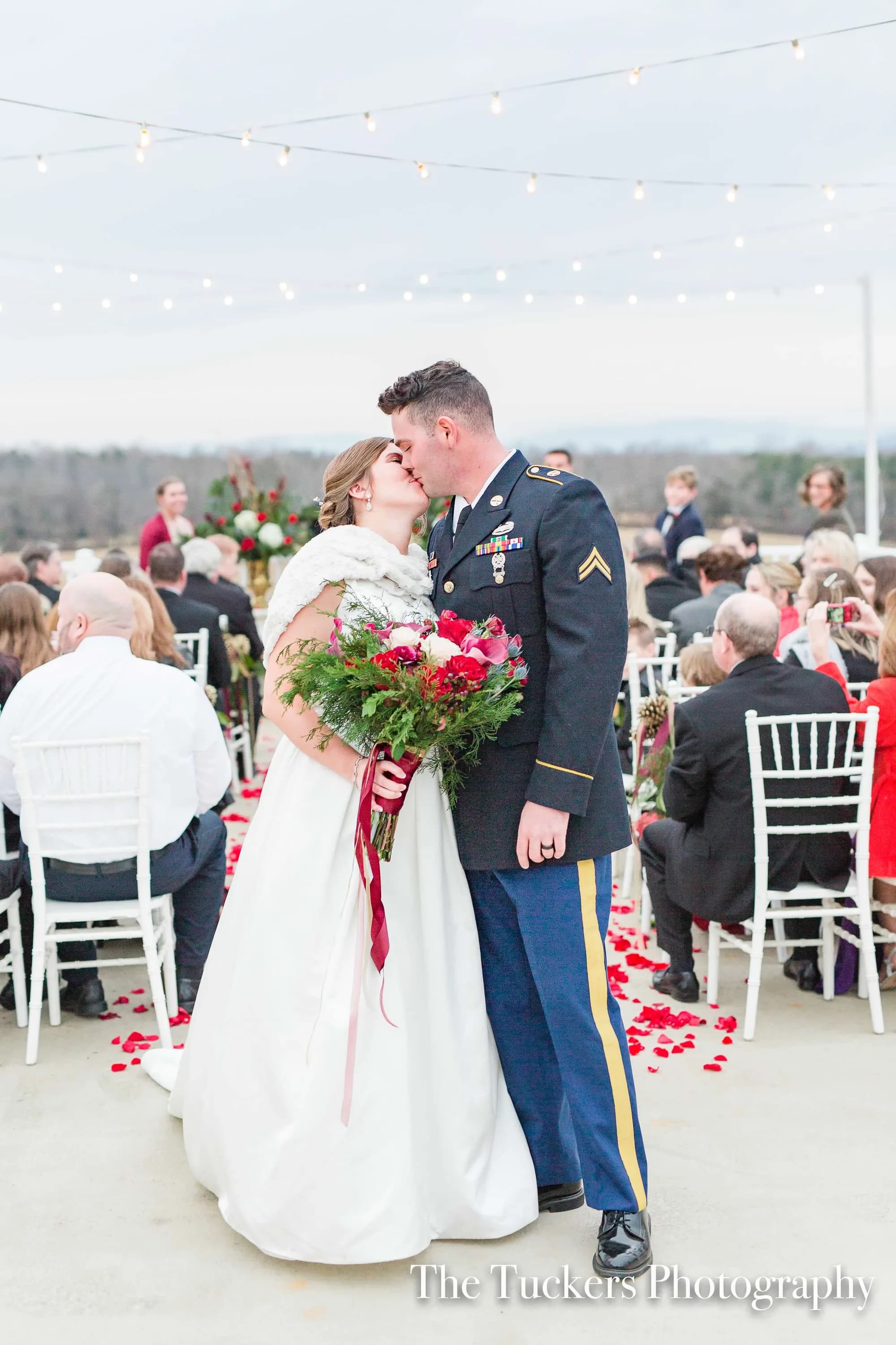 Military groom in dress uniform kisses bride at outdoor ceremony aisle lined with rose petals and string lights