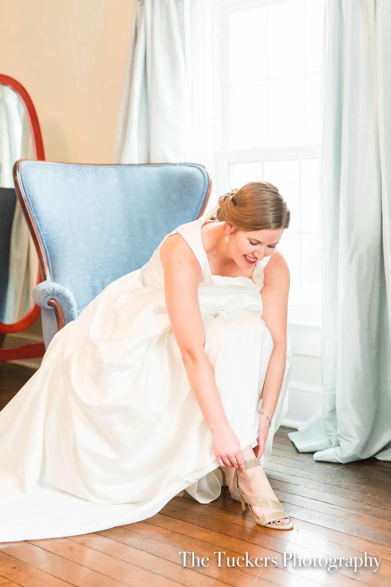 Smiling bride in white gown fastening her heels beside a blue velvet chair in a sunlit room