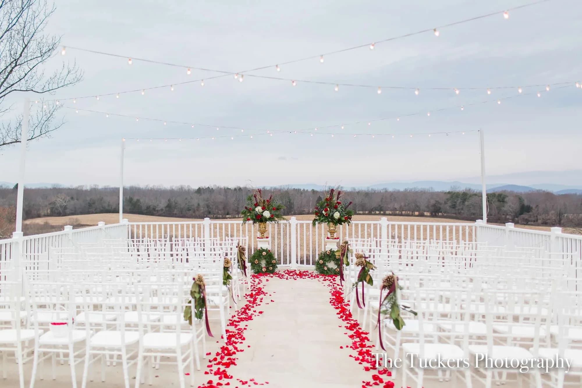 Outdoor ceremony aisle with red rose petals, white chairs, floral arches, and string lights overlooking Virginia mountains