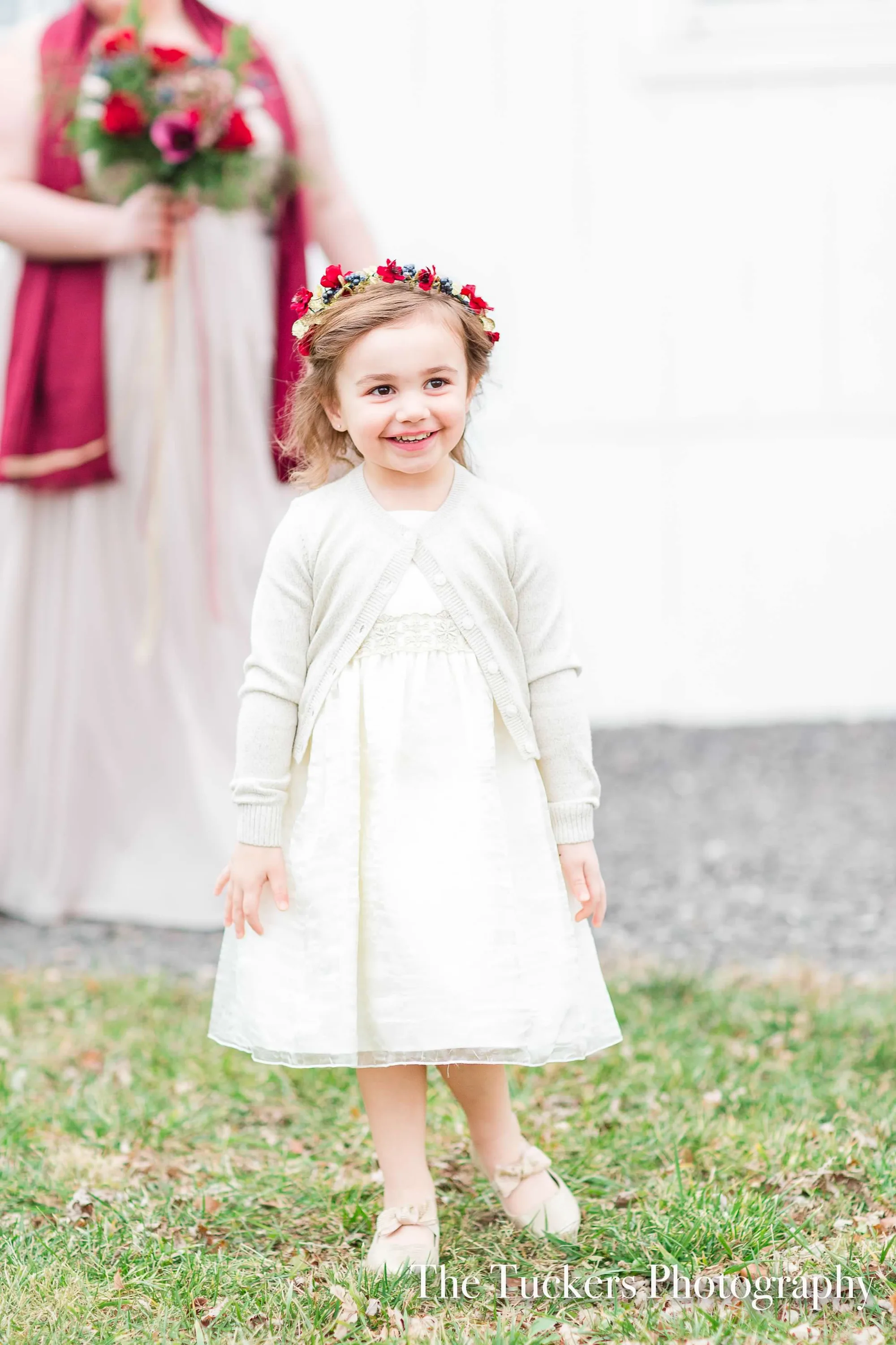 Smiling flower girl in white dress and red floral crown stands on lawn at a wedding