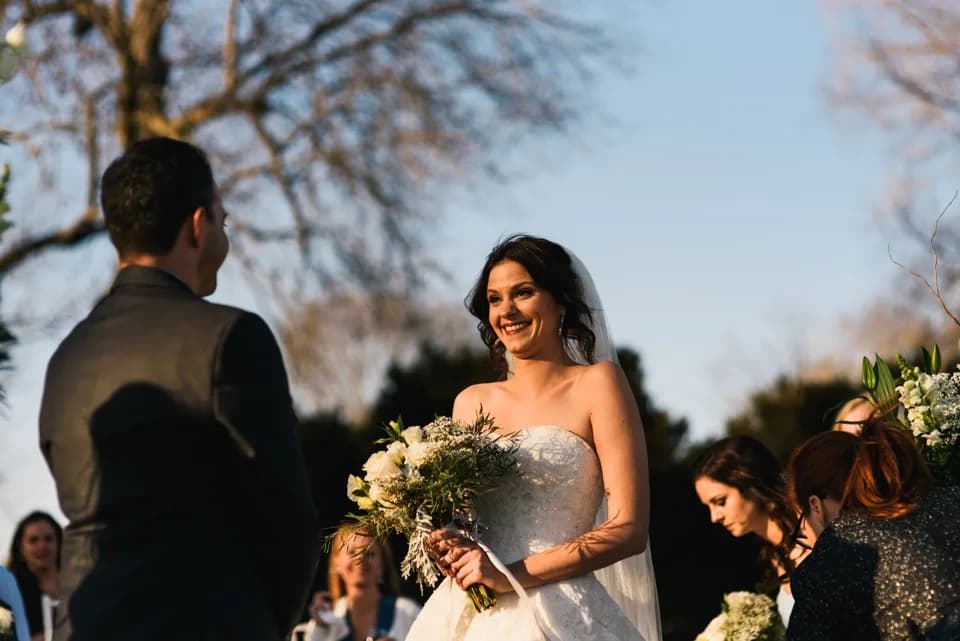Radiant bride smiling at groom during outdoor wedding ceremony, holding white bouquet in golden afternoon light