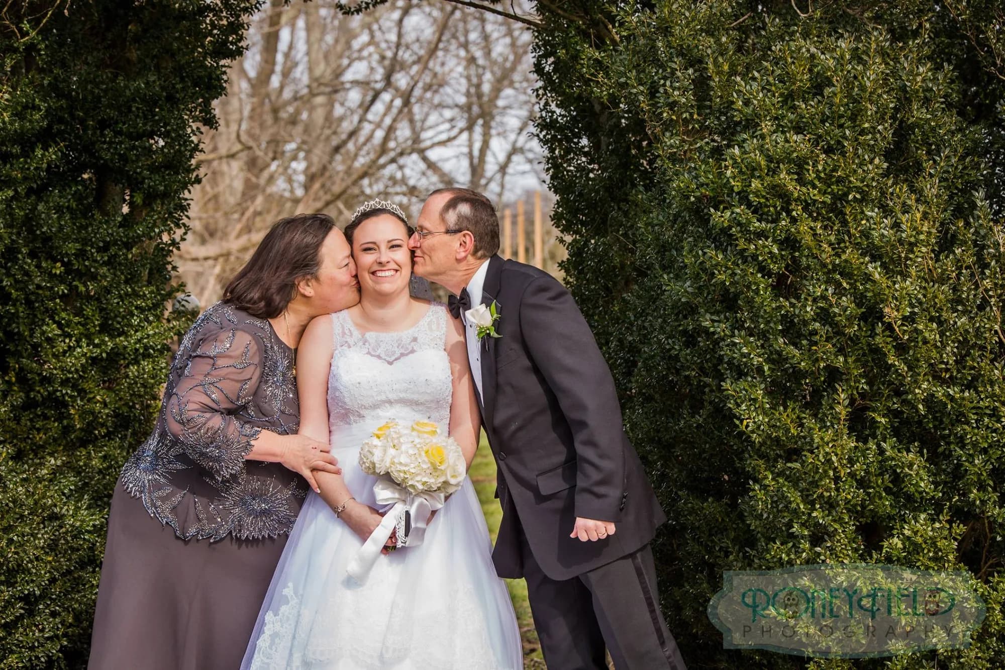 Bride smiles as both parents kiss her cheeks simultaneously in a garden archway on her wedding day