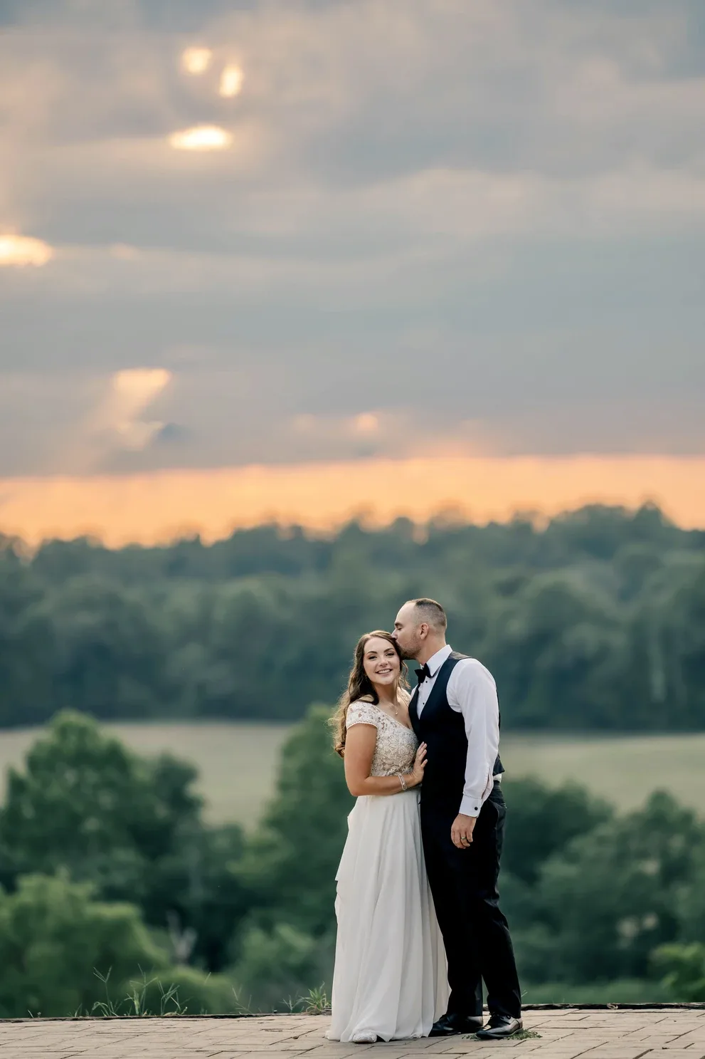 Groom kisses bride's cheek on Rixey Manor terrace at golden sunset over rolling Virginia countryside