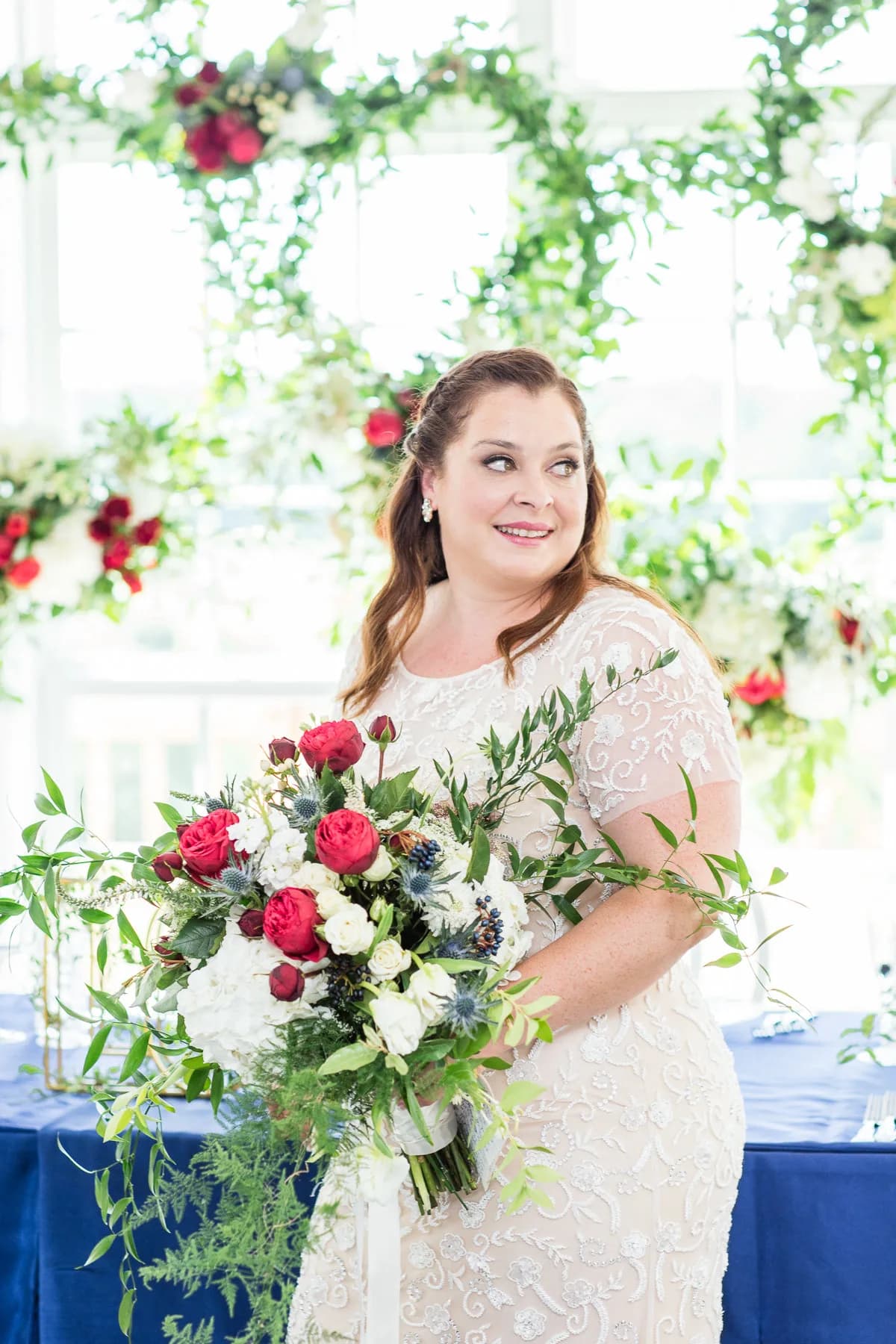 Smiling bride in lace gown holding a lush red and white bouquet before a floral arch backdrop