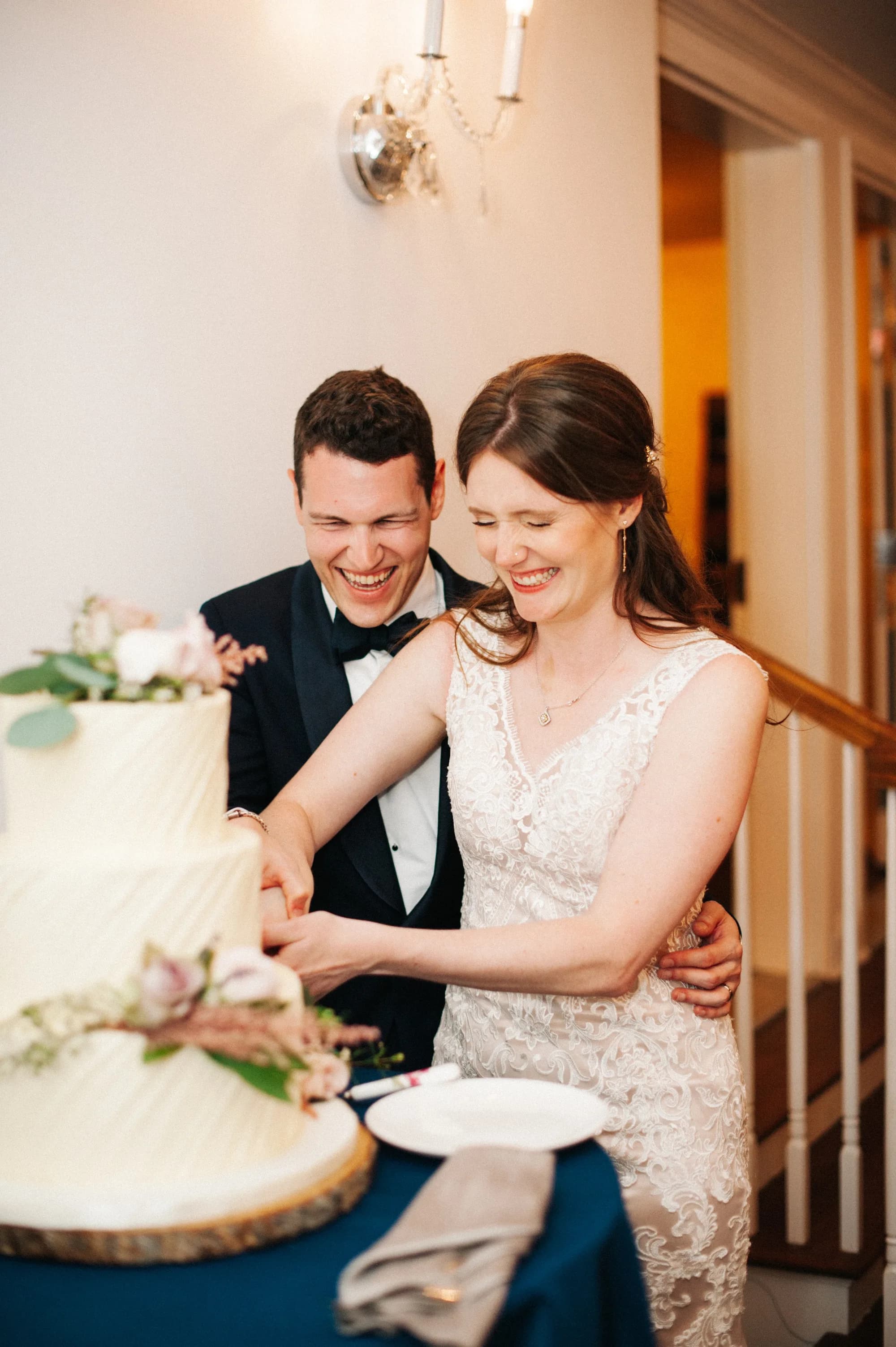 Bride and groom laugh joyfully while cutting their white floral wedding cake at their reception