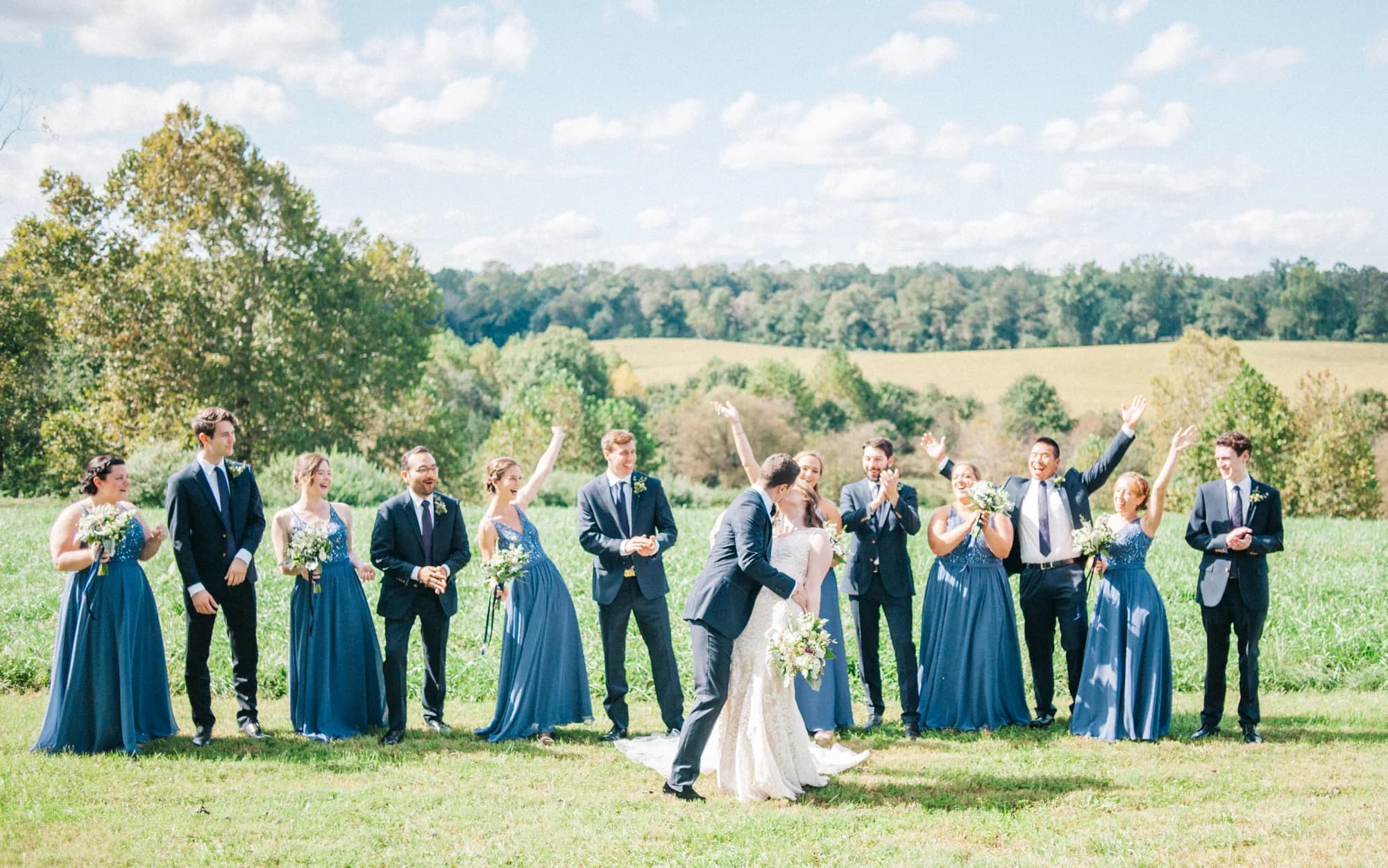 Bride and groom share a kiss while wedding party celebrates on Rixey Manor's sunlit green fields