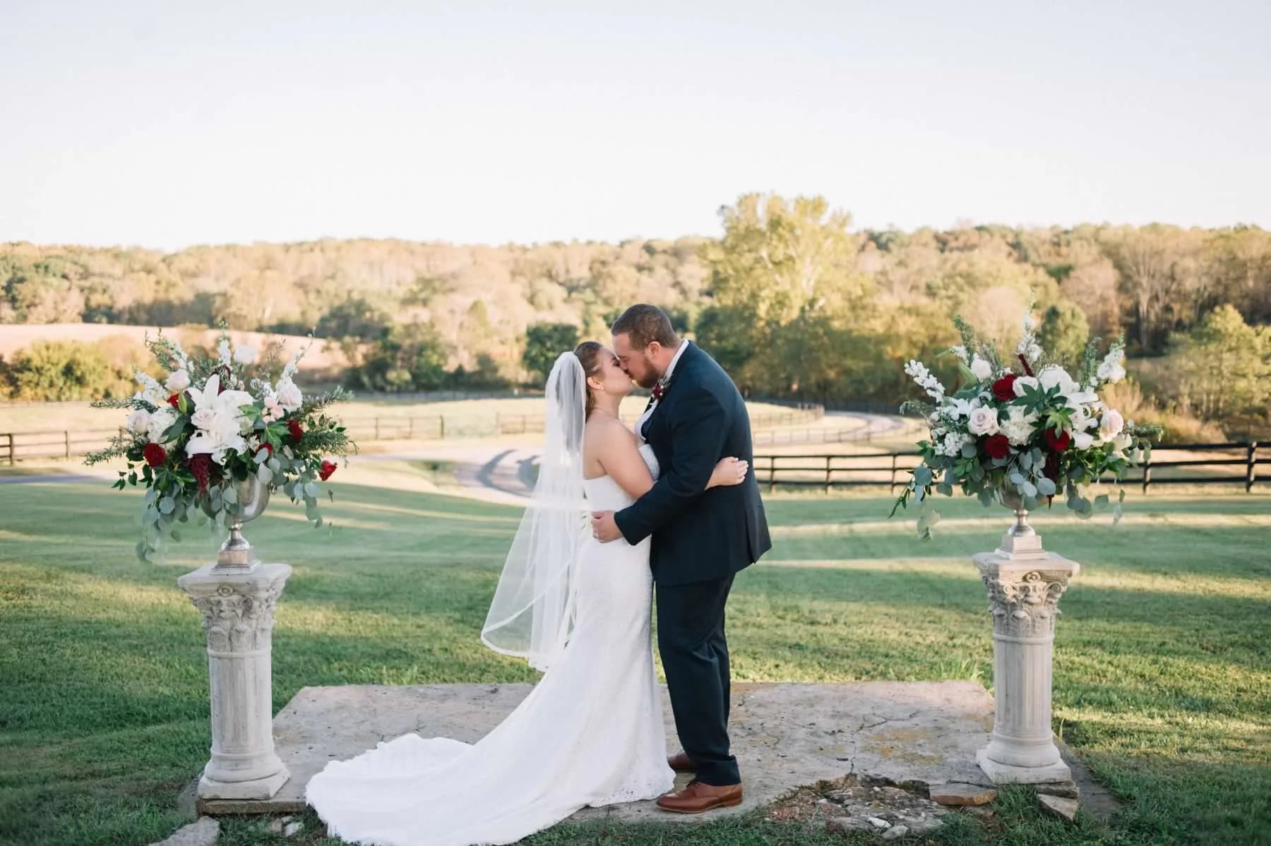 Bride and groom kiss at outdoor wedding ceremony with floral columns and pastoral landscape backdrop